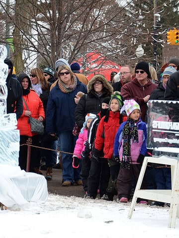 People in winter clothes stand in line outdoors, looking at an ice sculpture on a snowy day—a classic scene from winter festivals celebrated around the world.