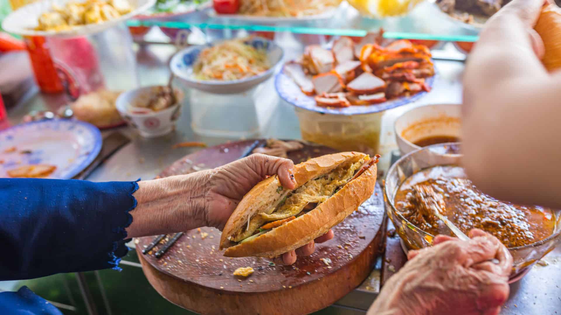 A person prepares a Vietnamese banh mi sandwich at a vibrant street food stall.