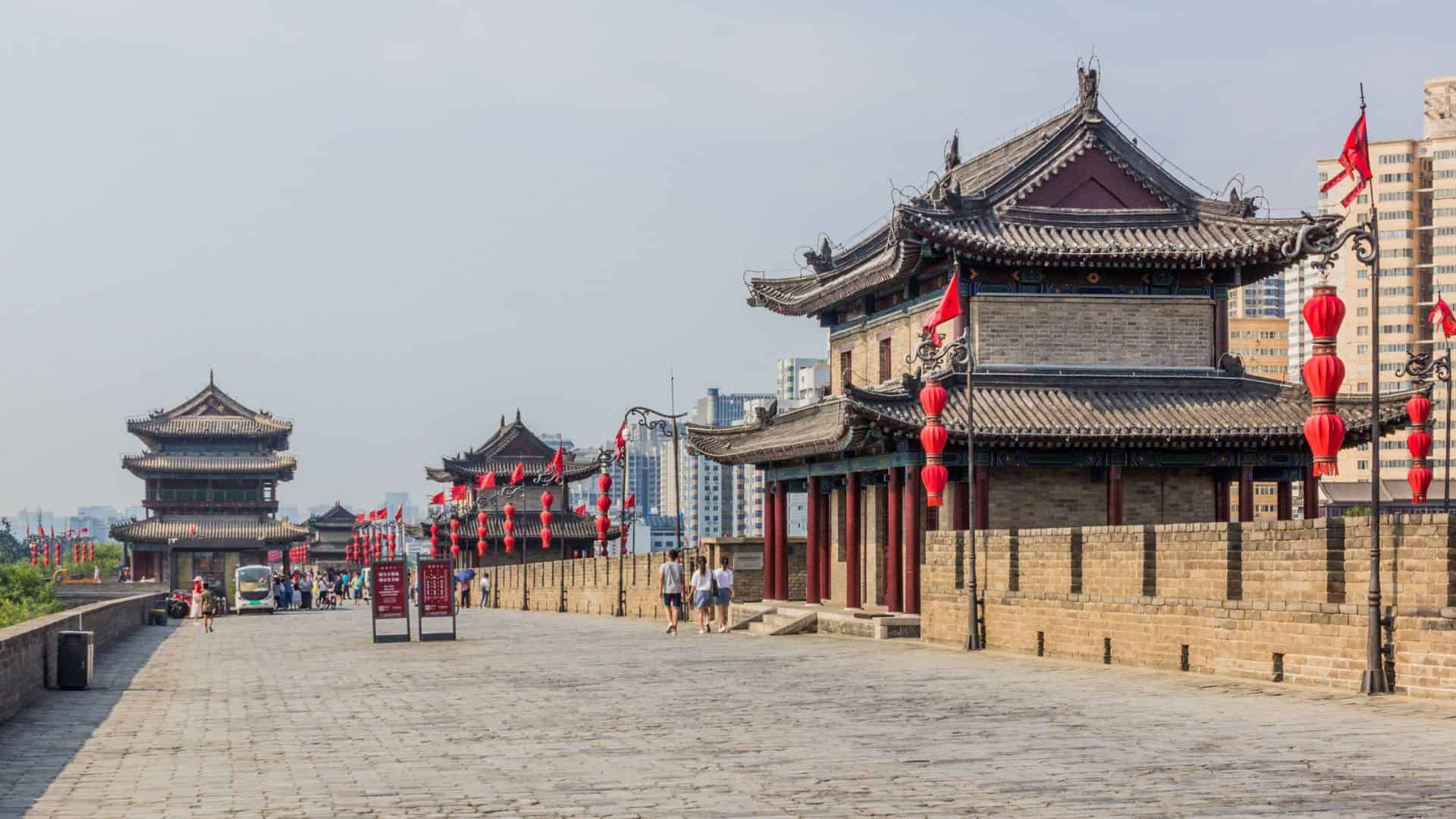 Ancient city wall with traditional Chinese architecture and red lanterns, people walking, city buildings in background.