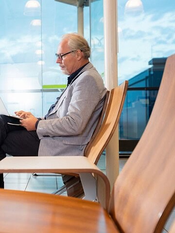 Older man in business attire working on a laptop in an airport lounge with his suitcase beside him.