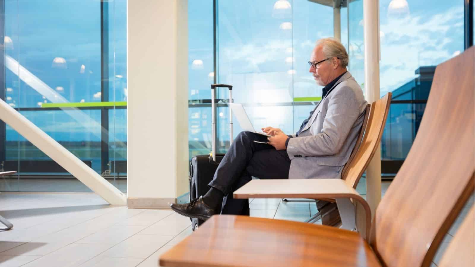 Older man in business attire working on a laptop in an airport lounge with his suitcase beside him.