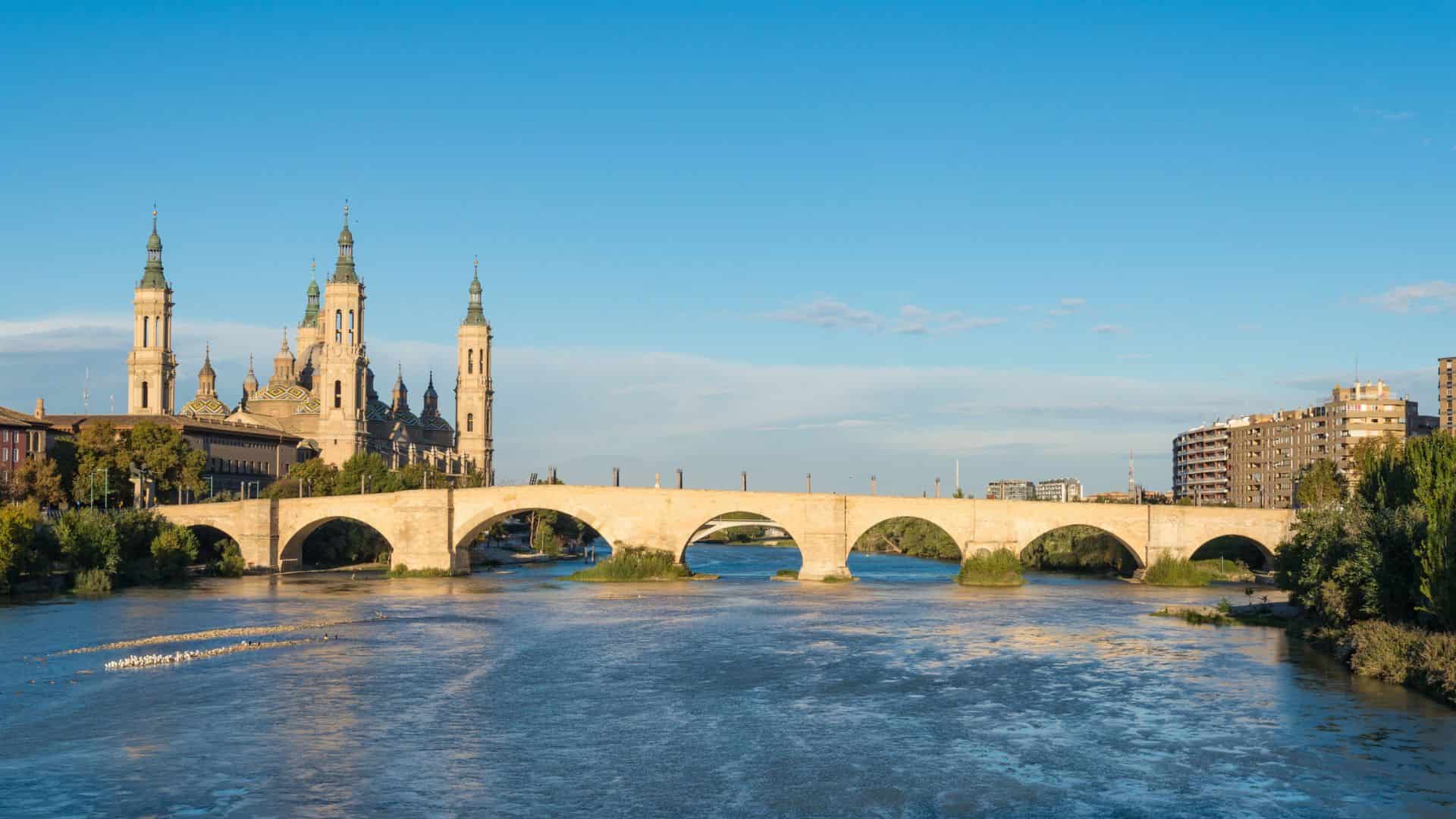 Stone bridge over a wide river, with a large cathedral and city buildings in the background under a clear sky.