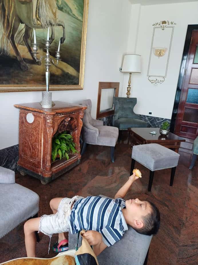 A young boy in a striped shirt lounges on a couch, eating, in an ornate room with classic furniture and artwork—capturing the relaxed charm of 4 days in Castel Gandolfo.