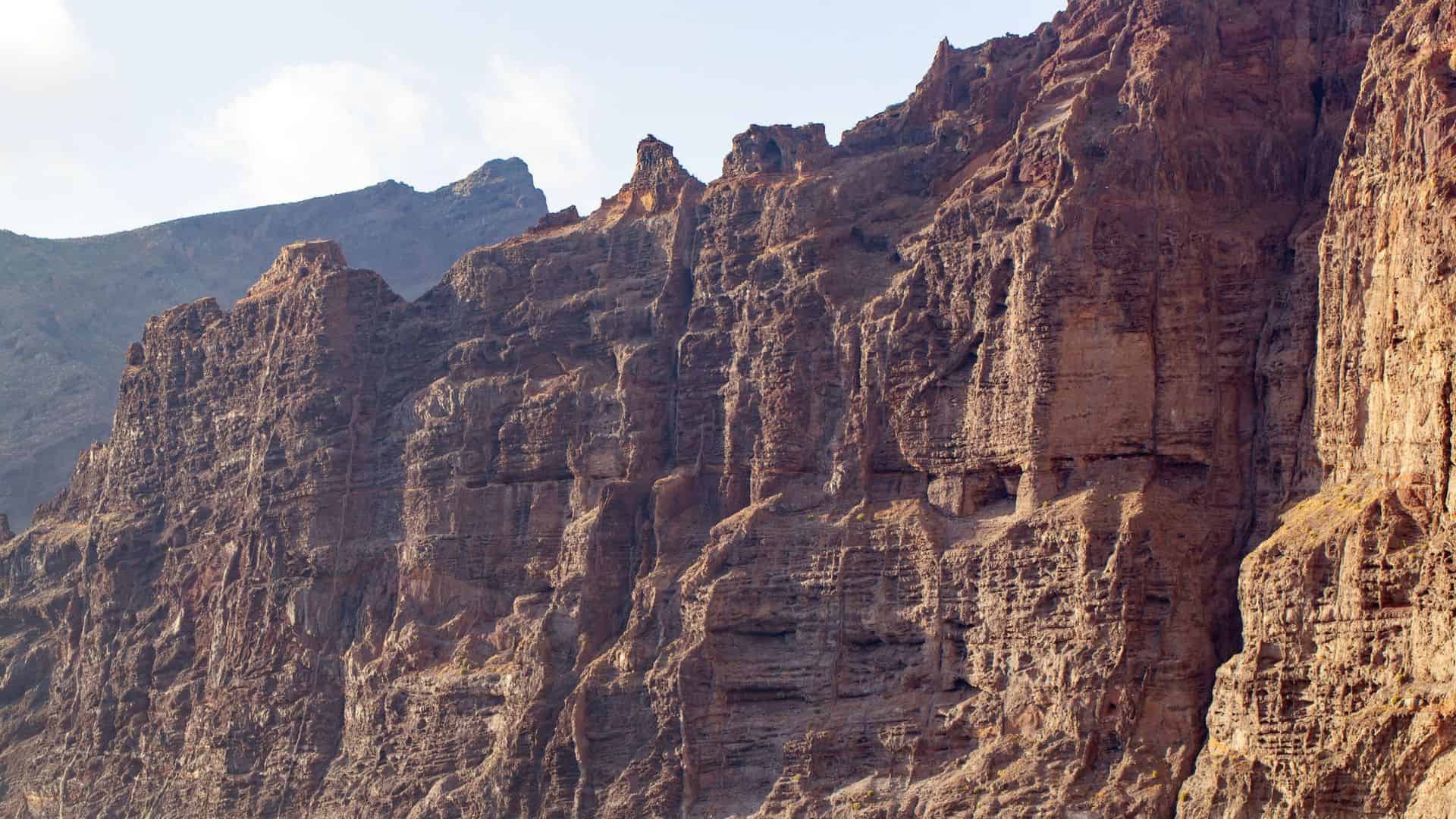 Sheer, rugged cliffs of brown rock rise steeply against a partly cloudy sky.