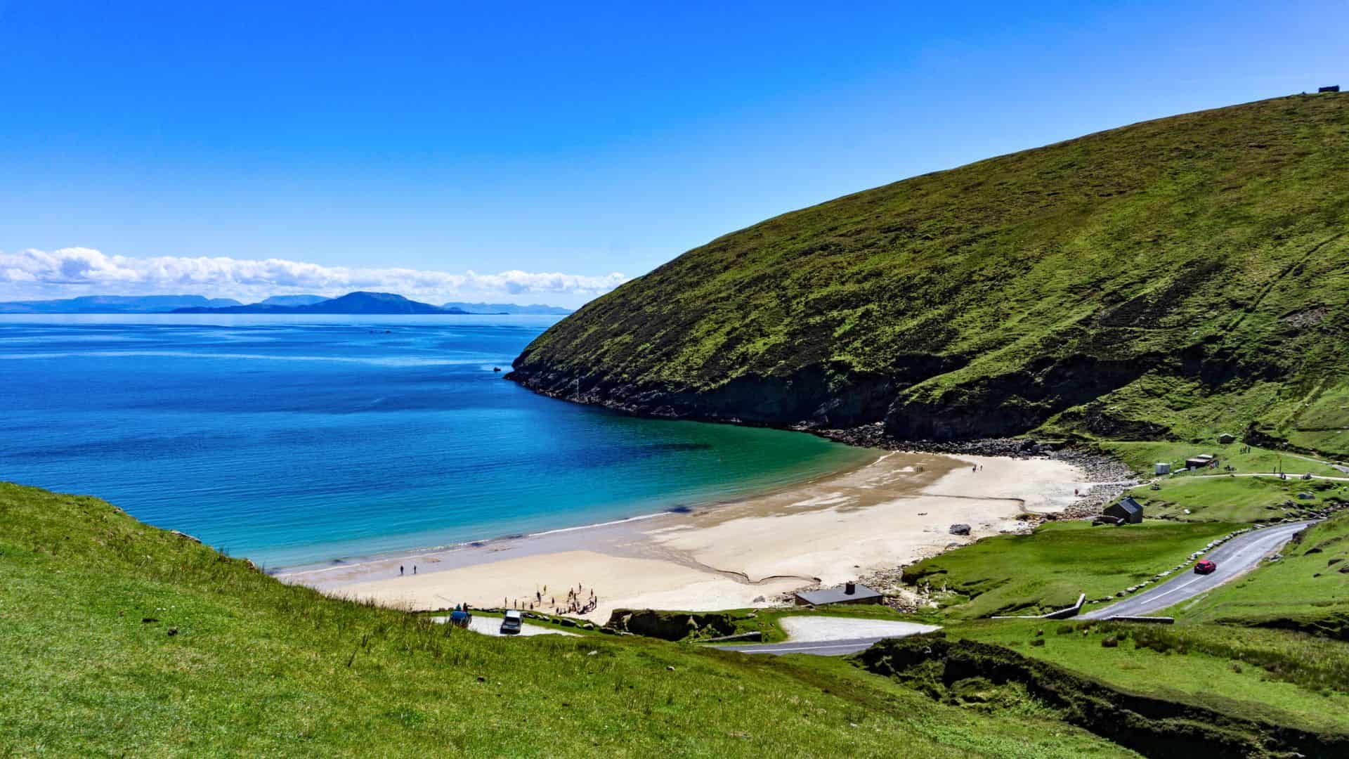 Sandy beach with turquoise water, green hills, and a winding road under a clear blue sky.