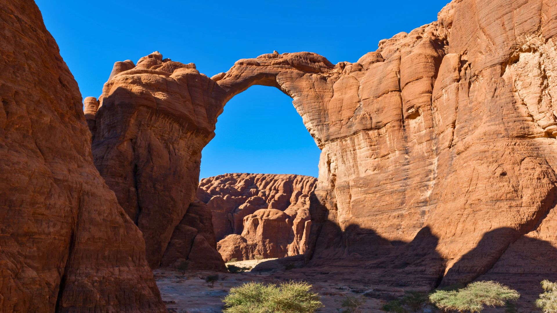 A natural stone arch rises between tall red cliffs under a bright blue sky in a desert landscape.
