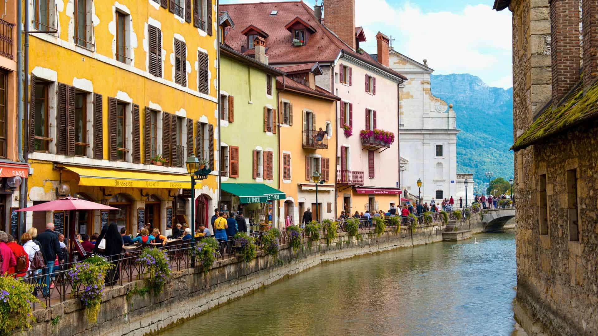Colorful buildings and outdoor cafes line a canal in a charming European town with mountains in the background.
