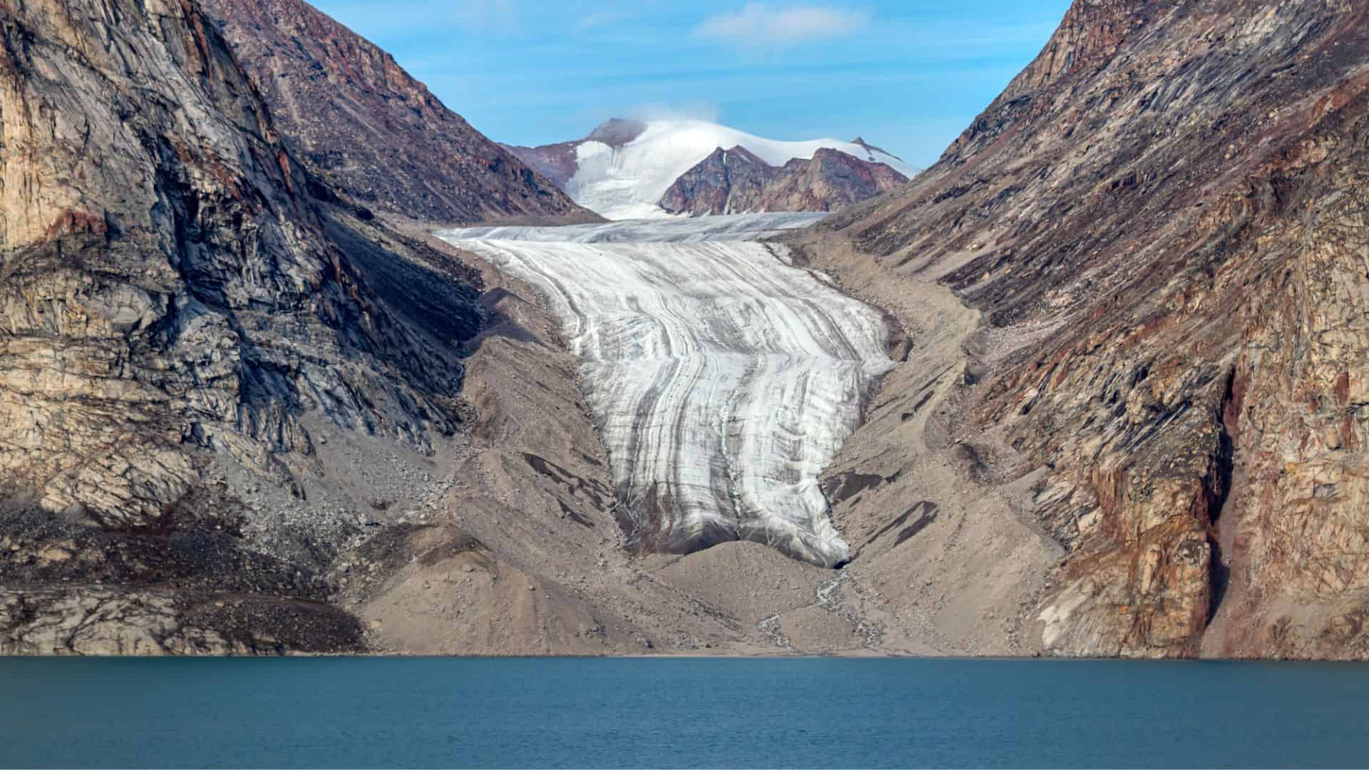 A glacier flows down a rocky mountain valley toward a blue lake under a clear sky.