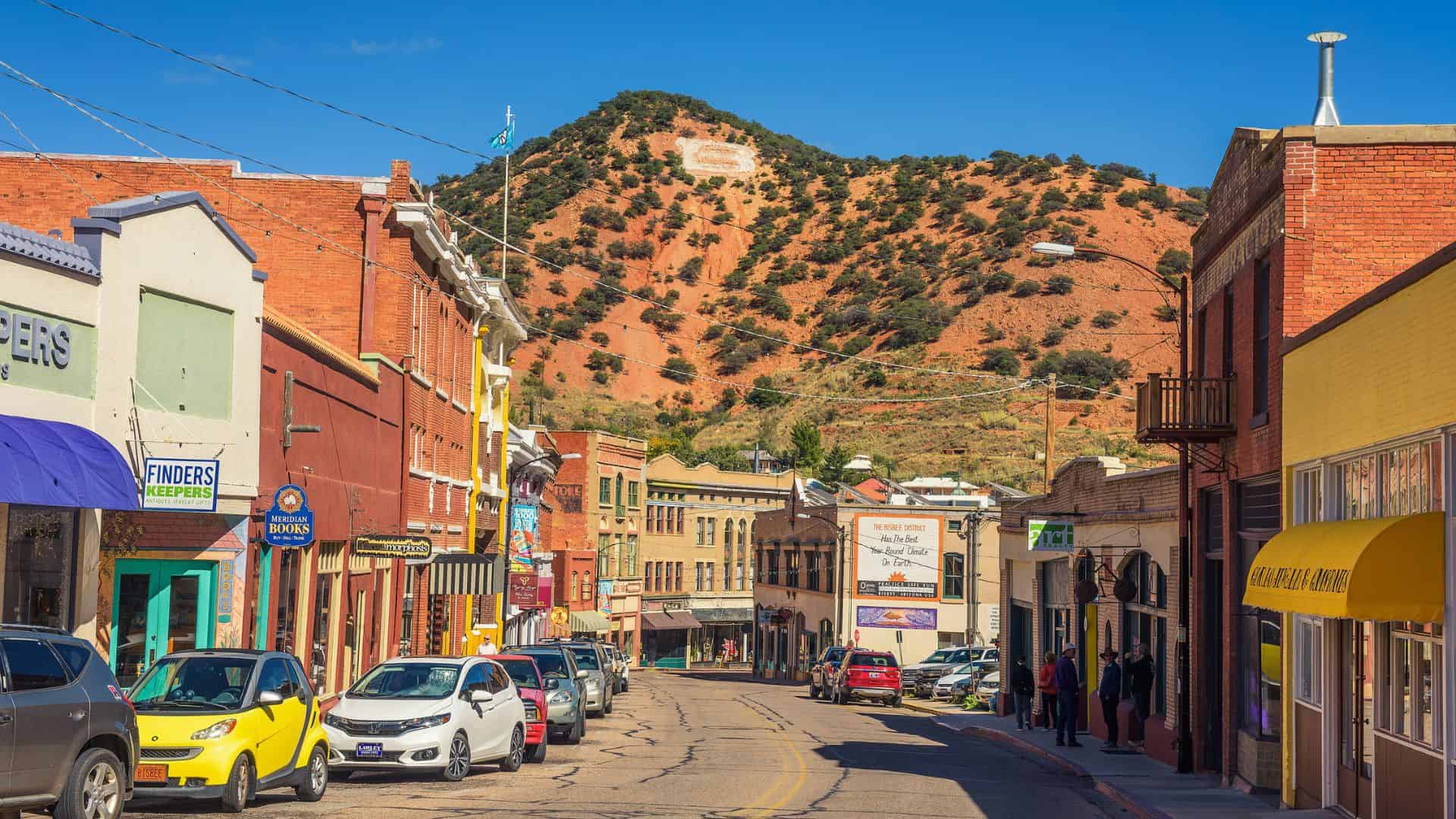 Colorful shops and parked cars line a sunny street with a rocky hill and a white "B" in the background.