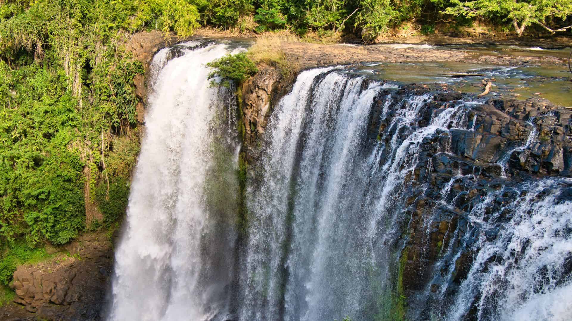 A wide waterfall cascades over rocky cliffs surrounded by lush green trees.
