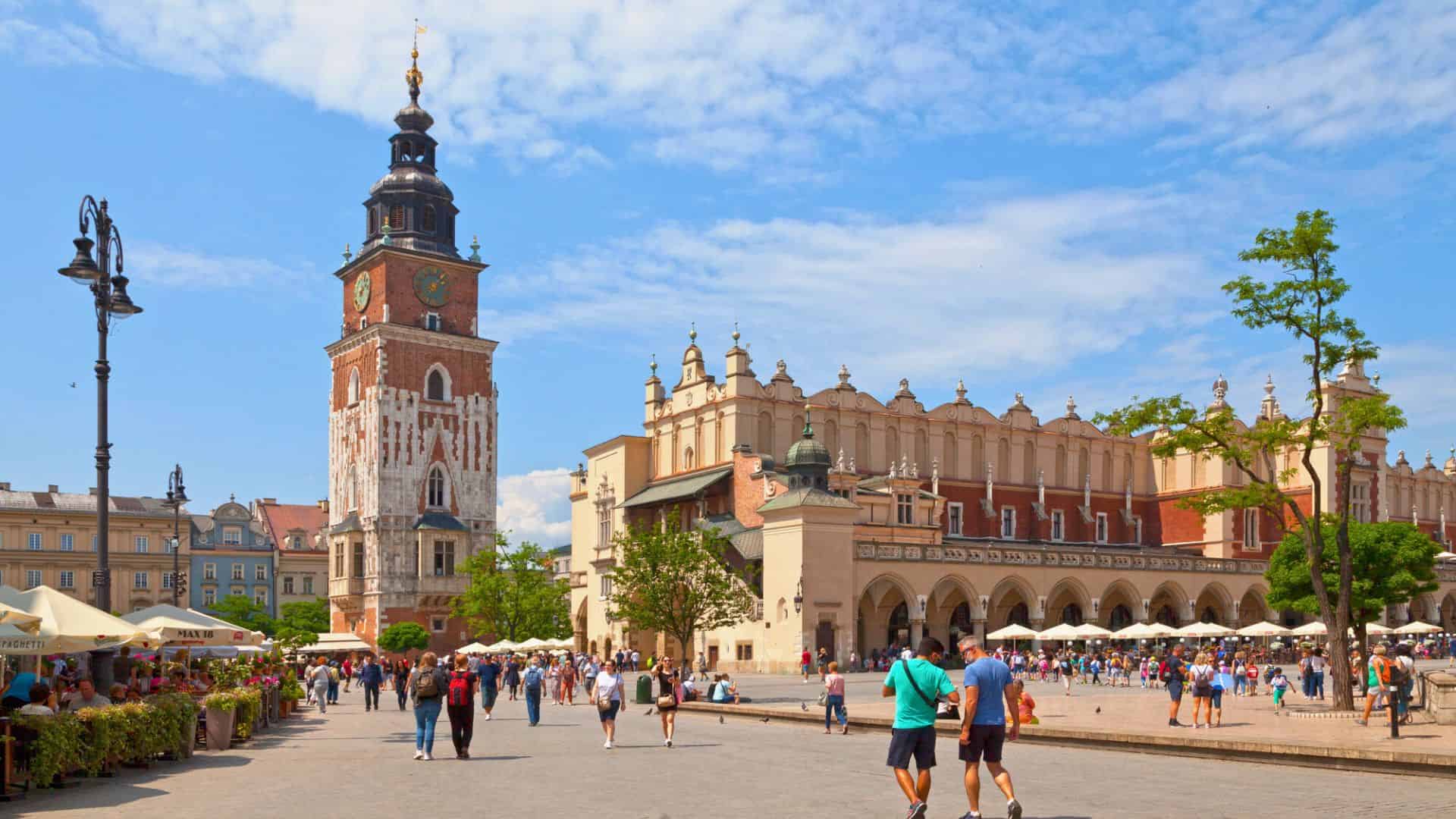 People walk in a sunny plaza with a historic tower and long arcade building in Kraków’s Main Market Square.