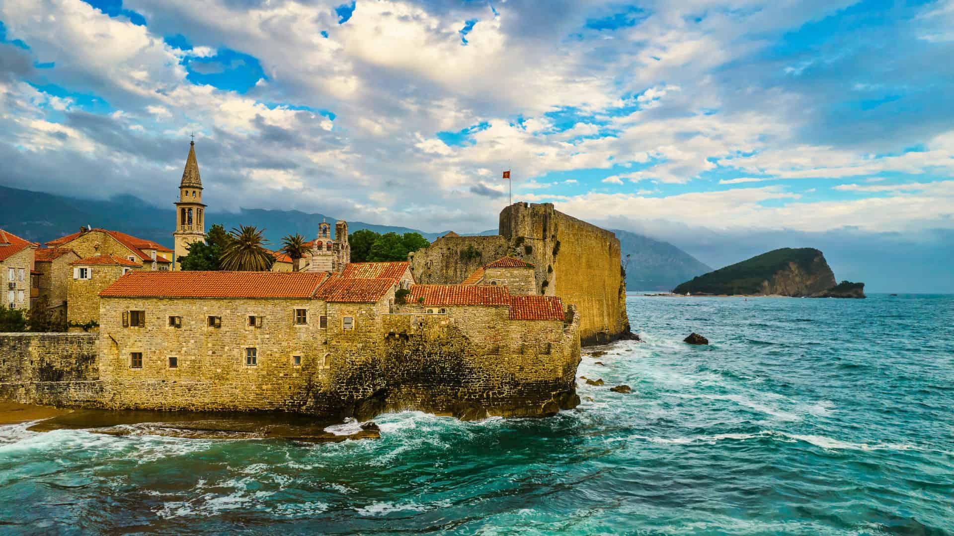 Old stone fortress with red rooftops by the sea, waves hitting walls, mountains and clouds in the background.