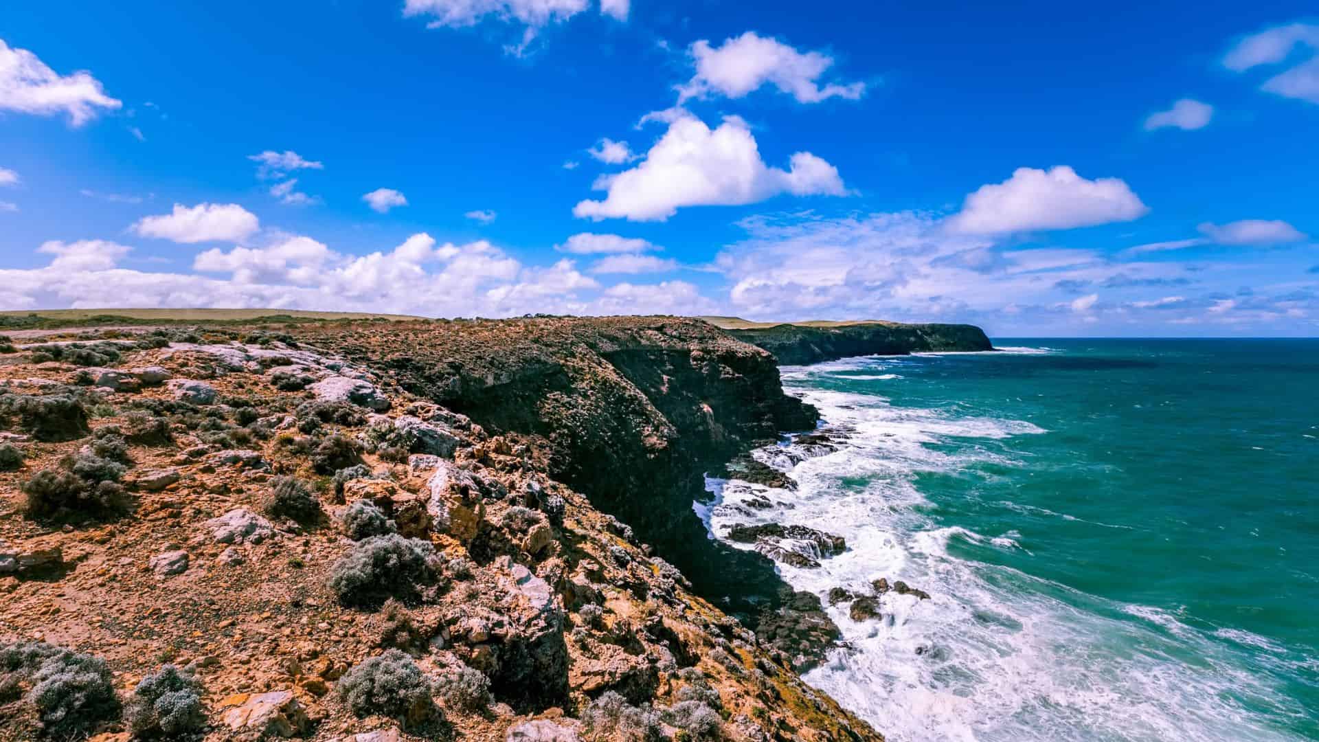 Rocky coastline with waves crashing against cliffs under a bright blue sky with scattered clouds.