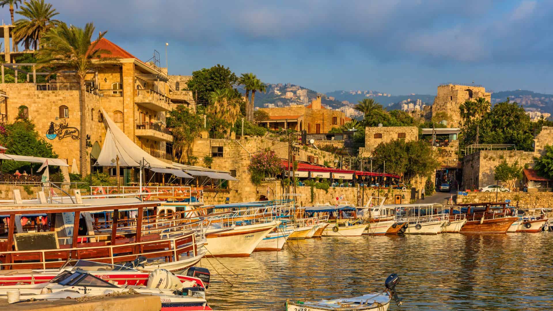 Colorful boats docked along a historic waterfront with stone buildings and palm trees under a blue sky.