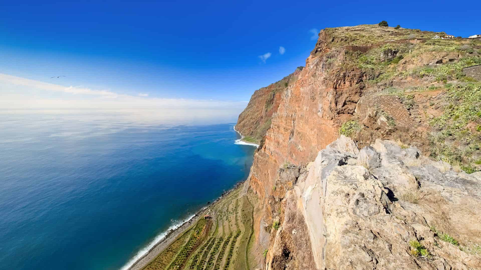 Steep red cliffs overlooking a blue ocean with terraced fields below under a clear sky.