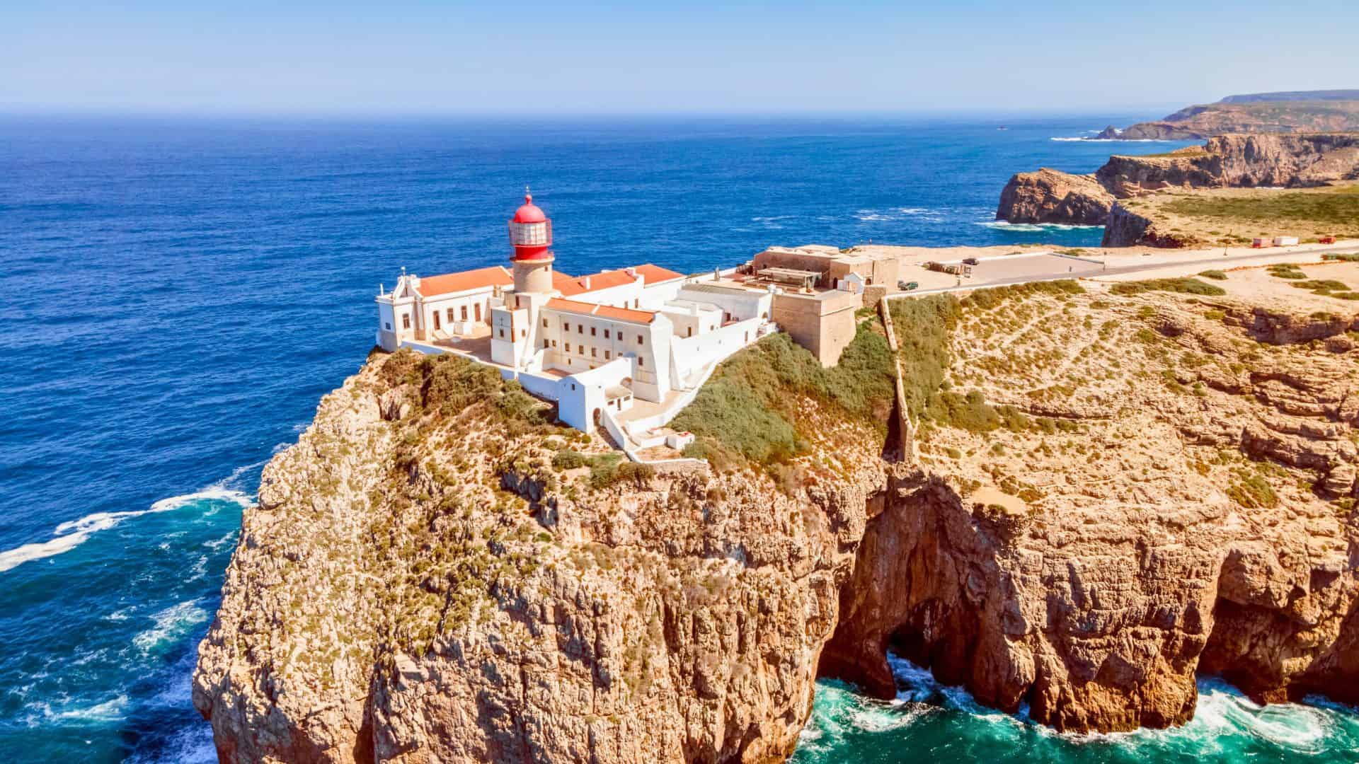Lighthouse with red dome on a cliff overlooking the ocean, surrounded by rugged coastline and blue water.