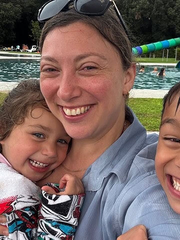 A woman and two smiling children pose for a close-up selfie by an outdoor pool with water slides at Parco della Gallinara.