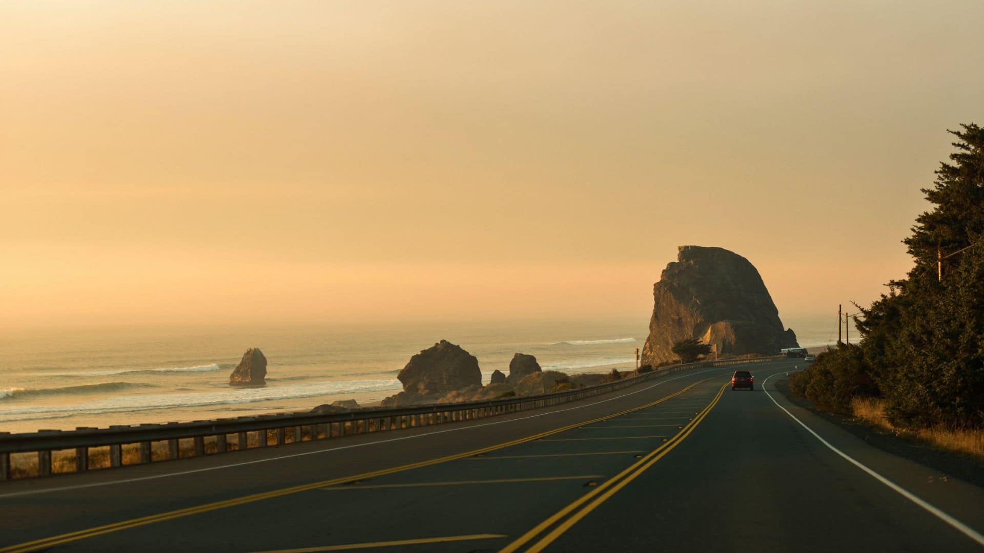 A coastal highway at sunset with rock formations by the ocean and a car driving along the road.