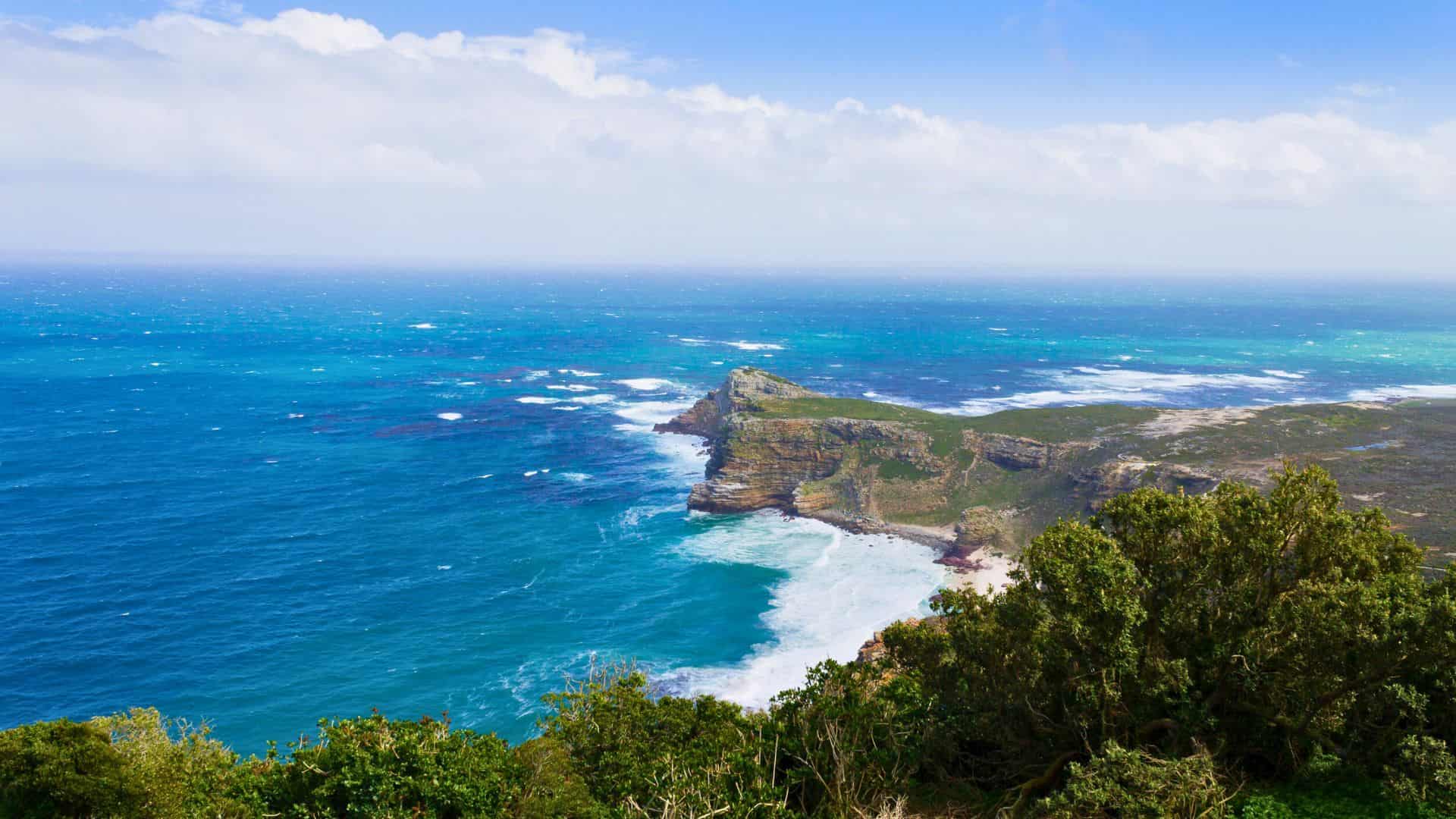 Rocky peninsula with waves crashing against cliffs, surrounded by blue ocean and lush green vegetation.