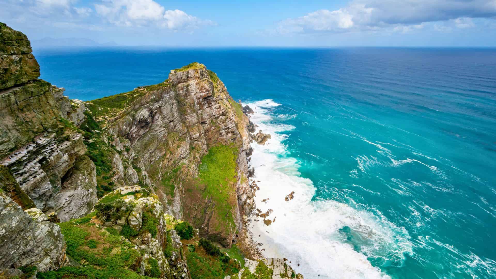 Steep rocky cliffs with green patches overlook turquoise ocean waves under a blue sky with scattered clouds.