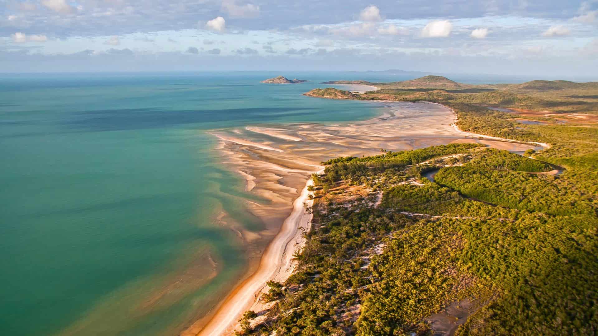 Aerial view of a sandy beach with turquoise water and green vegetation under a partly cloudy sky.