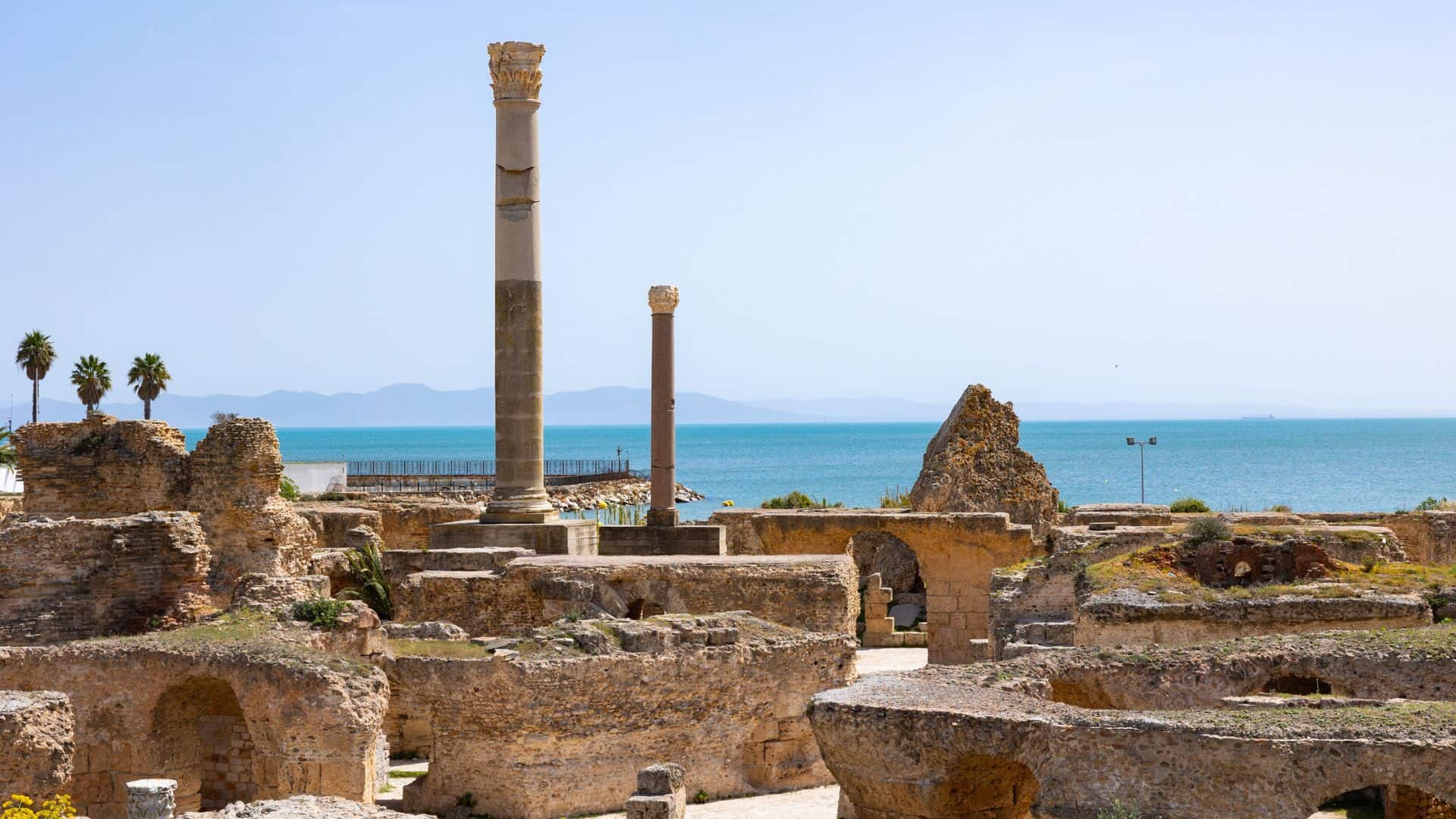 Ancient stone ruins with tall columns near the sea, with mountains and palm trees in the background.