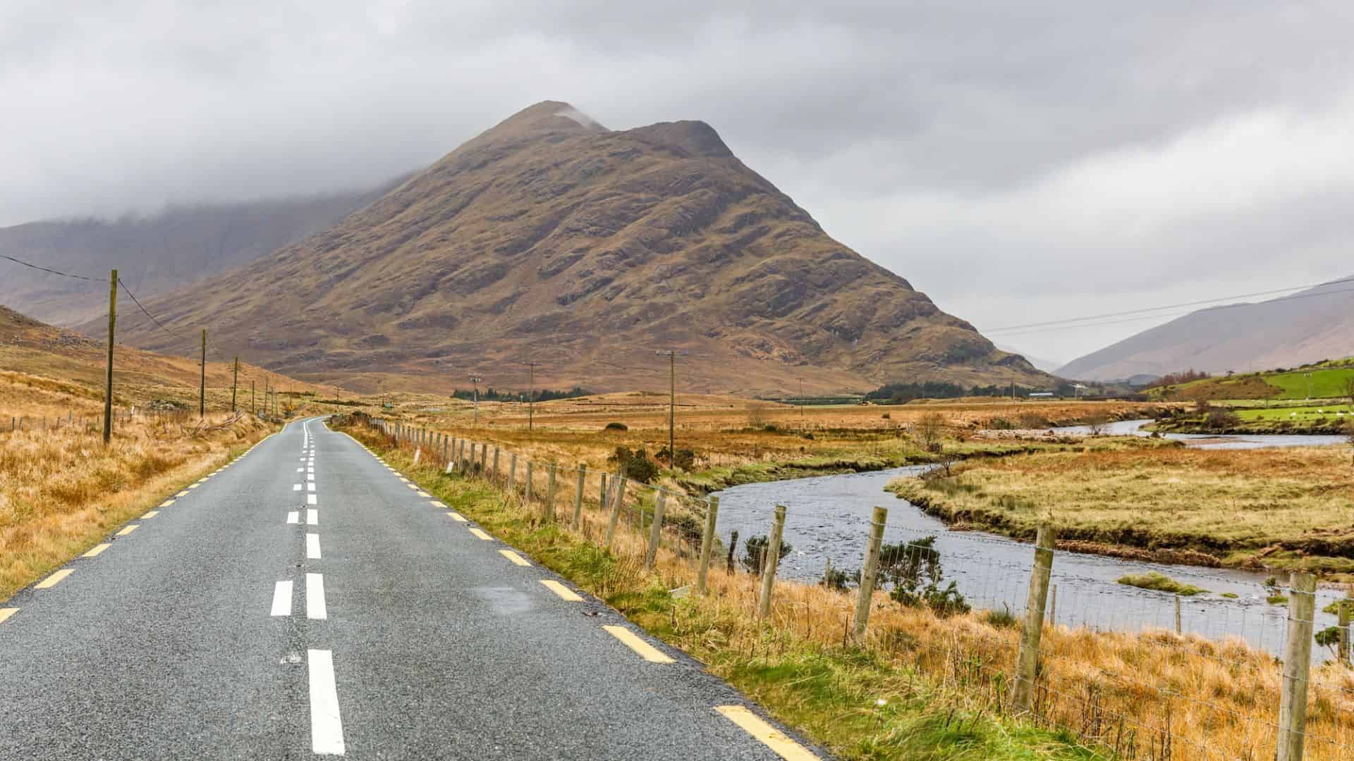 A rural road leads toward a mountain, with a river and grassy fields under a cloudy sky.