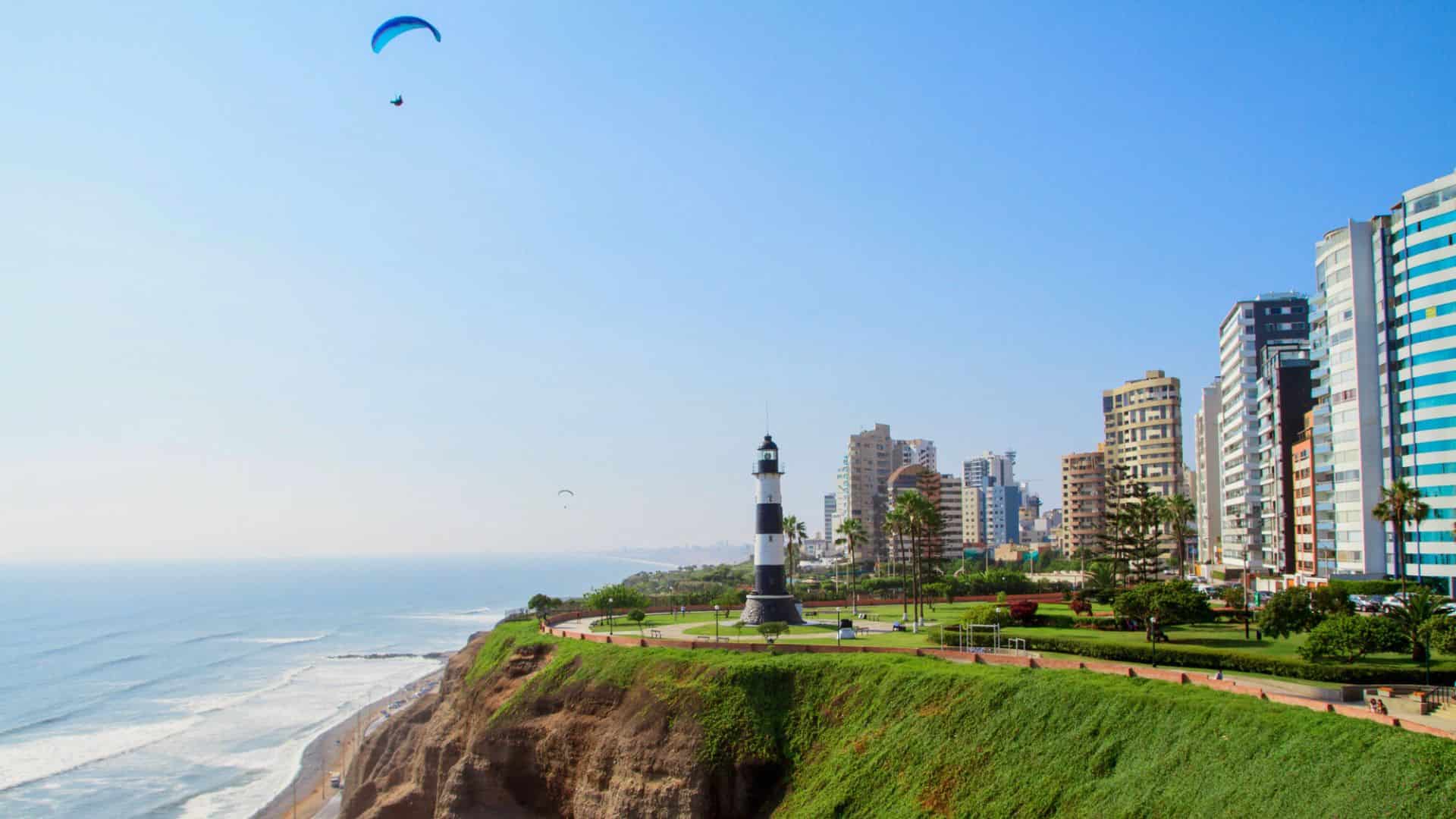A black-and-white lighthouse stands on a green cliff by the ocean, with tall buildings and a paraglider nearby.