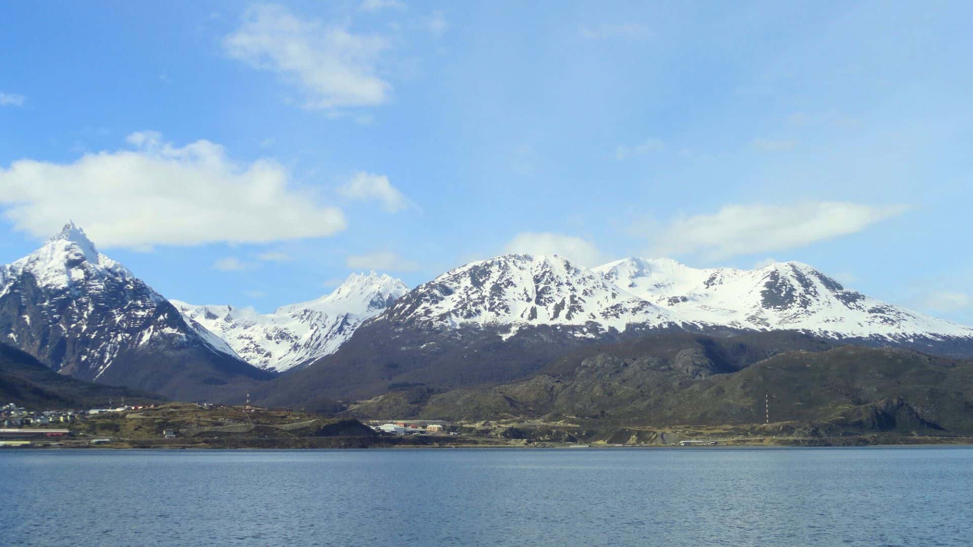 Snow-capped mountains under a blue sky with clouds, seen from across a calm body of water.