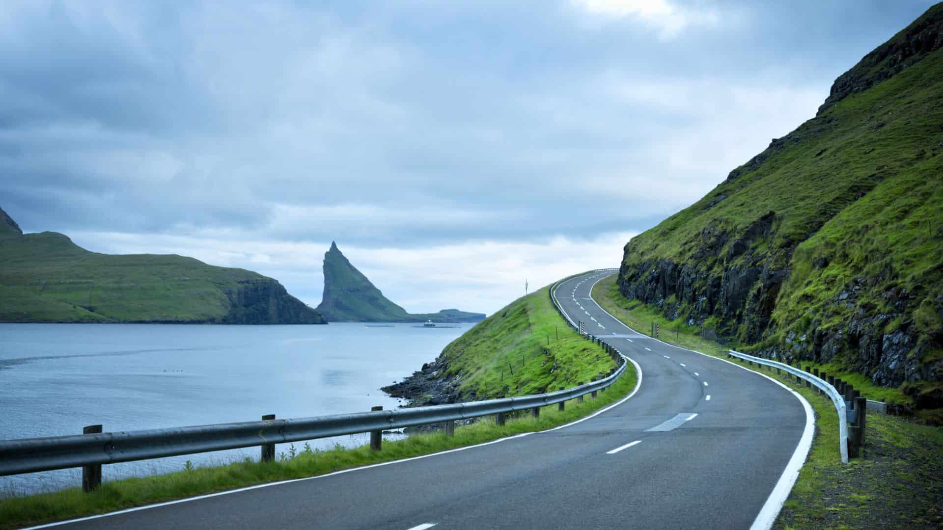 A winding coastal road curves along green hills beside a calm body of water under a cloudy sky.