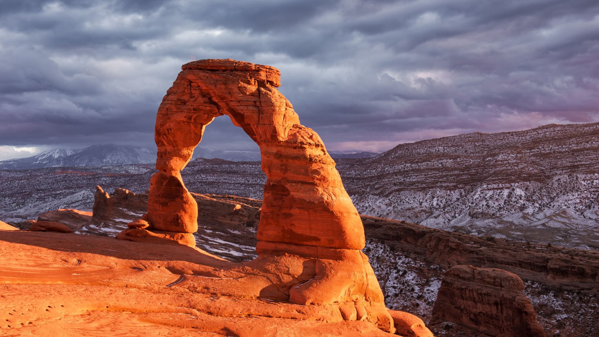 Delicate Arch in Utah glows in sunset light, with dramatic clouds and snowy mountains in the background.