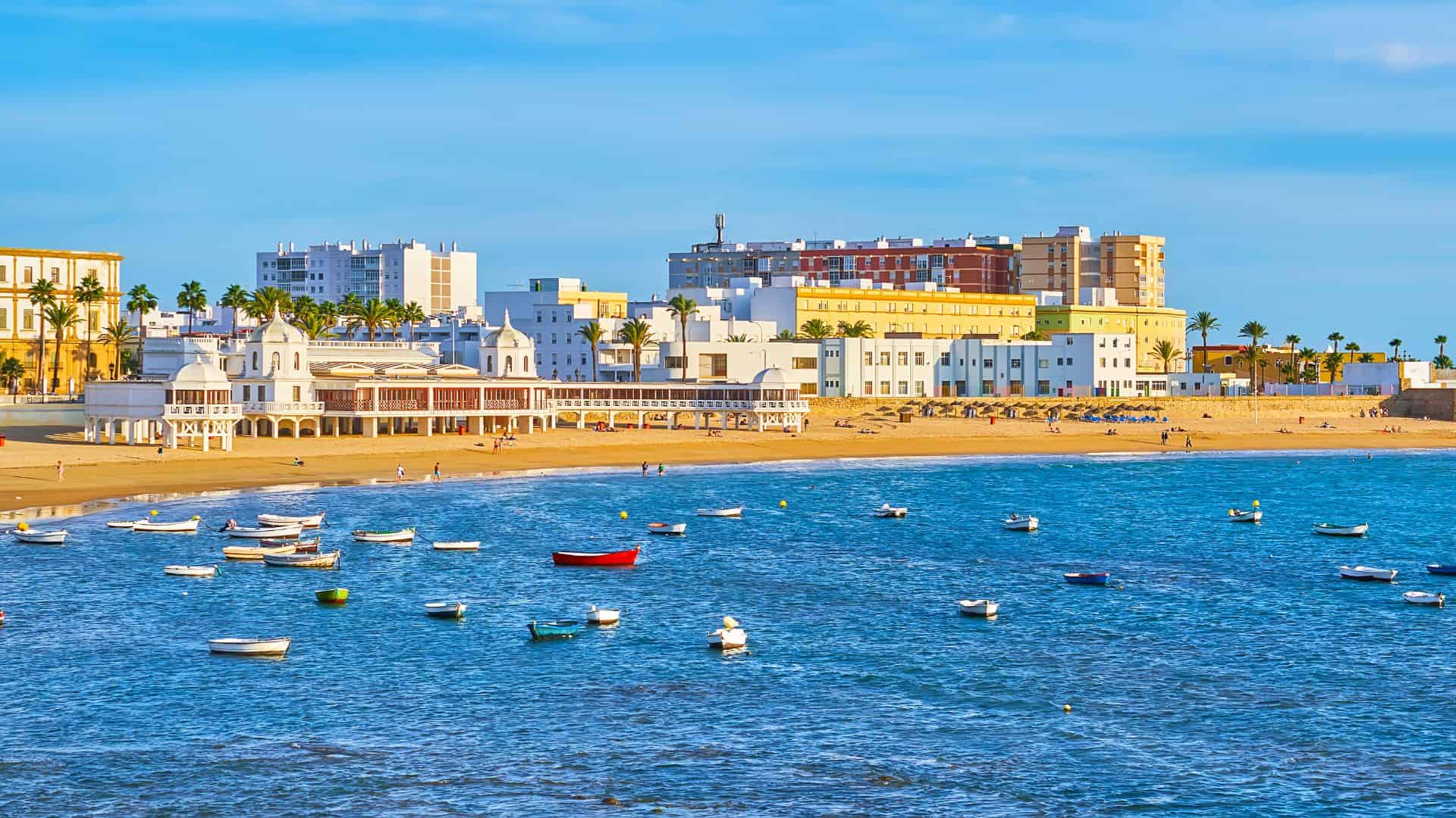 Boats float on blue water near a sandy beach and historic buildings under a sunny sky.