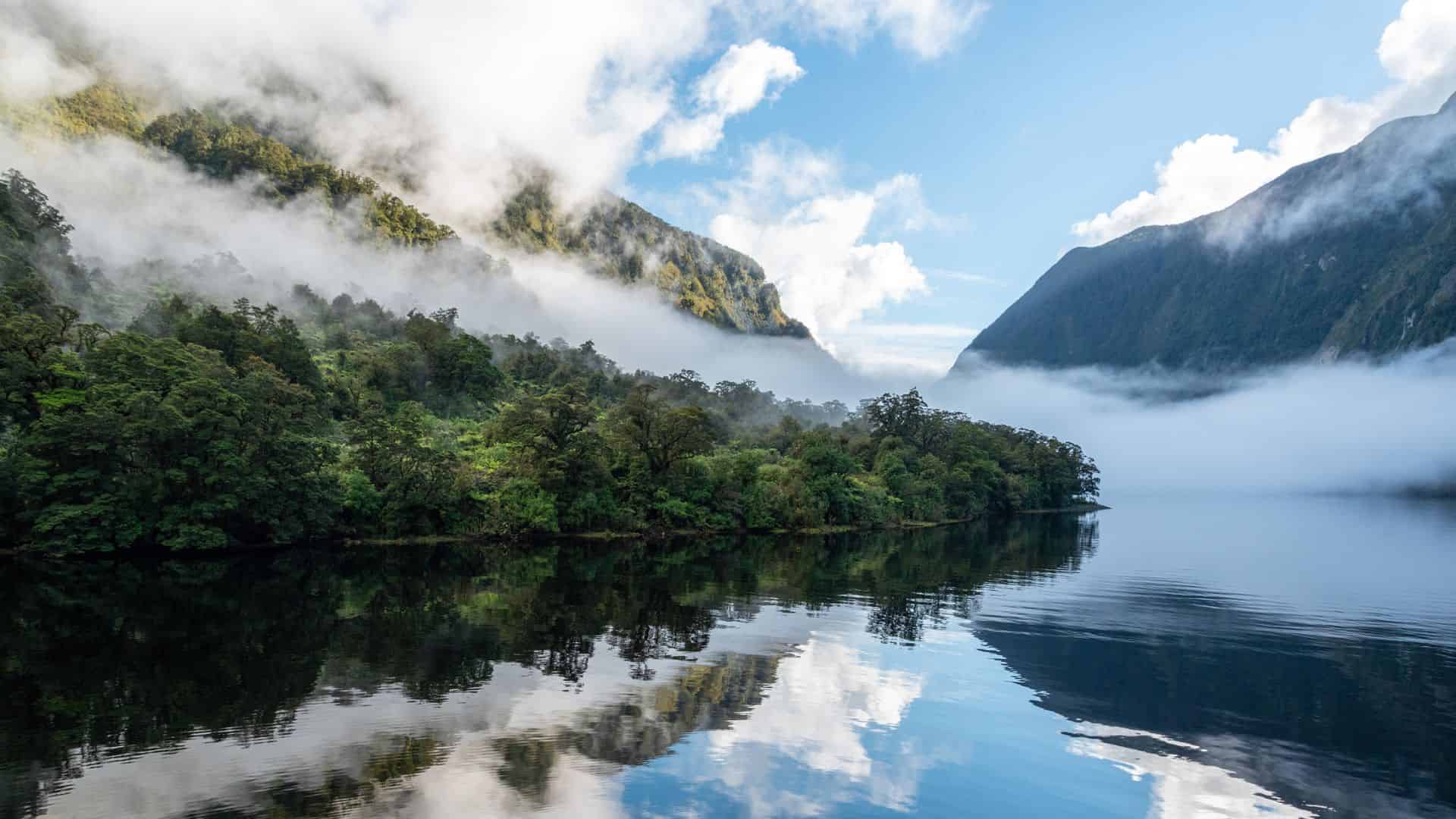 Clouds and mist drift over green forested mountains reflected in a calm, clear lake under a blue sky.
