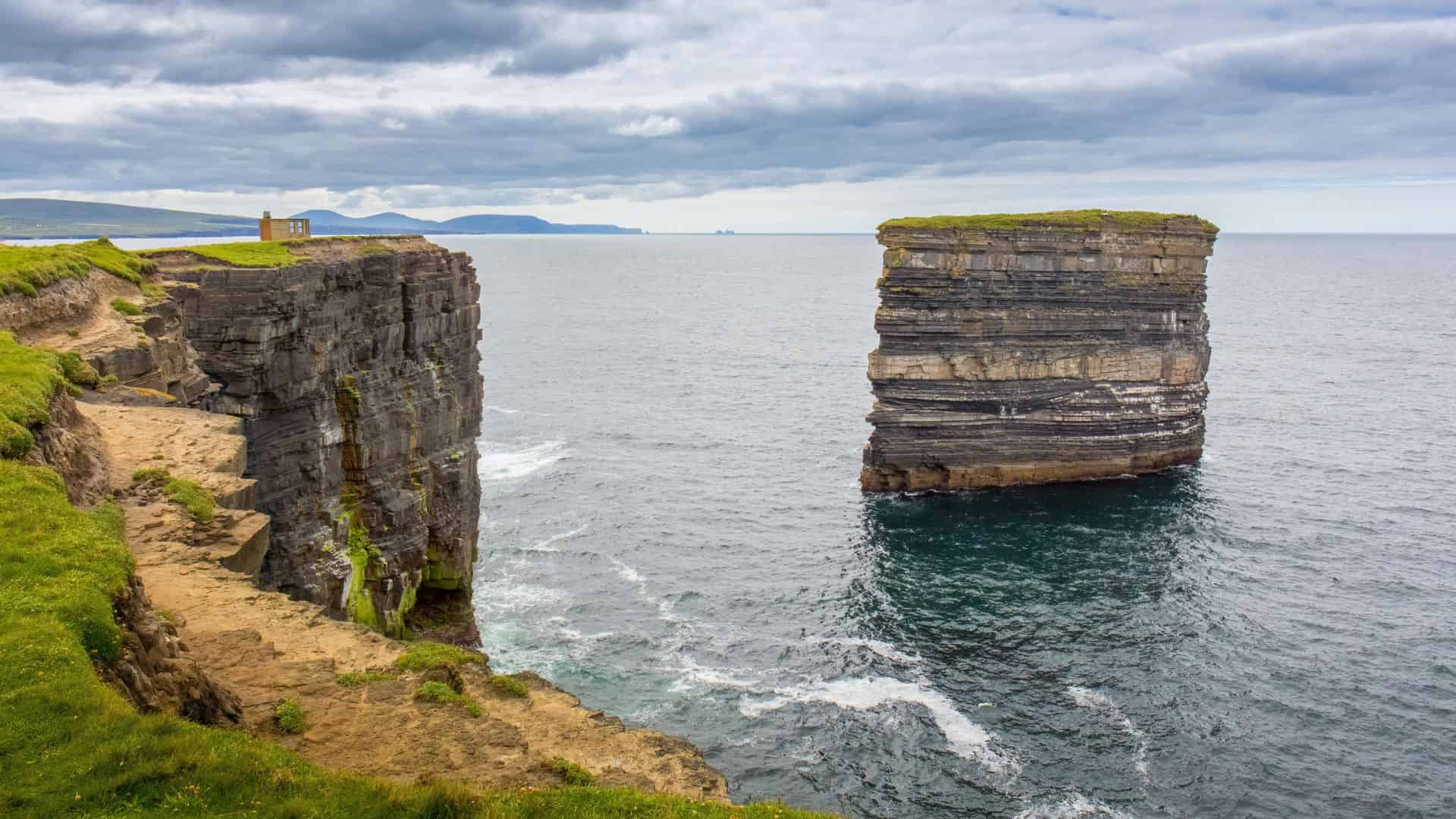A tall sea stack stands beside dramatic coastal cliffs under a cloudy sky, with waves crashing below.