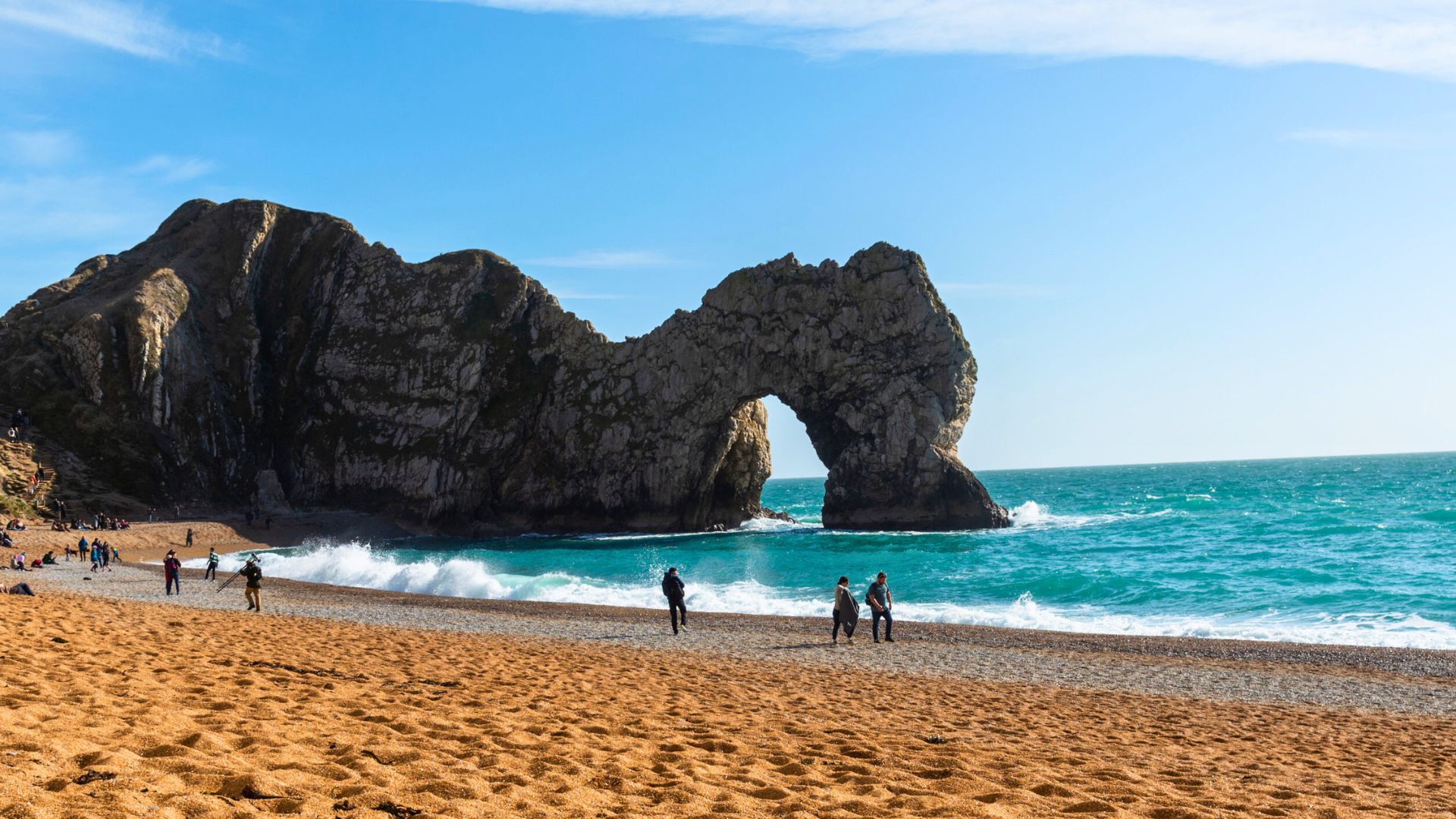 People walk on a sandy beach near a large rock arch by turquoise sea under a blue sky.