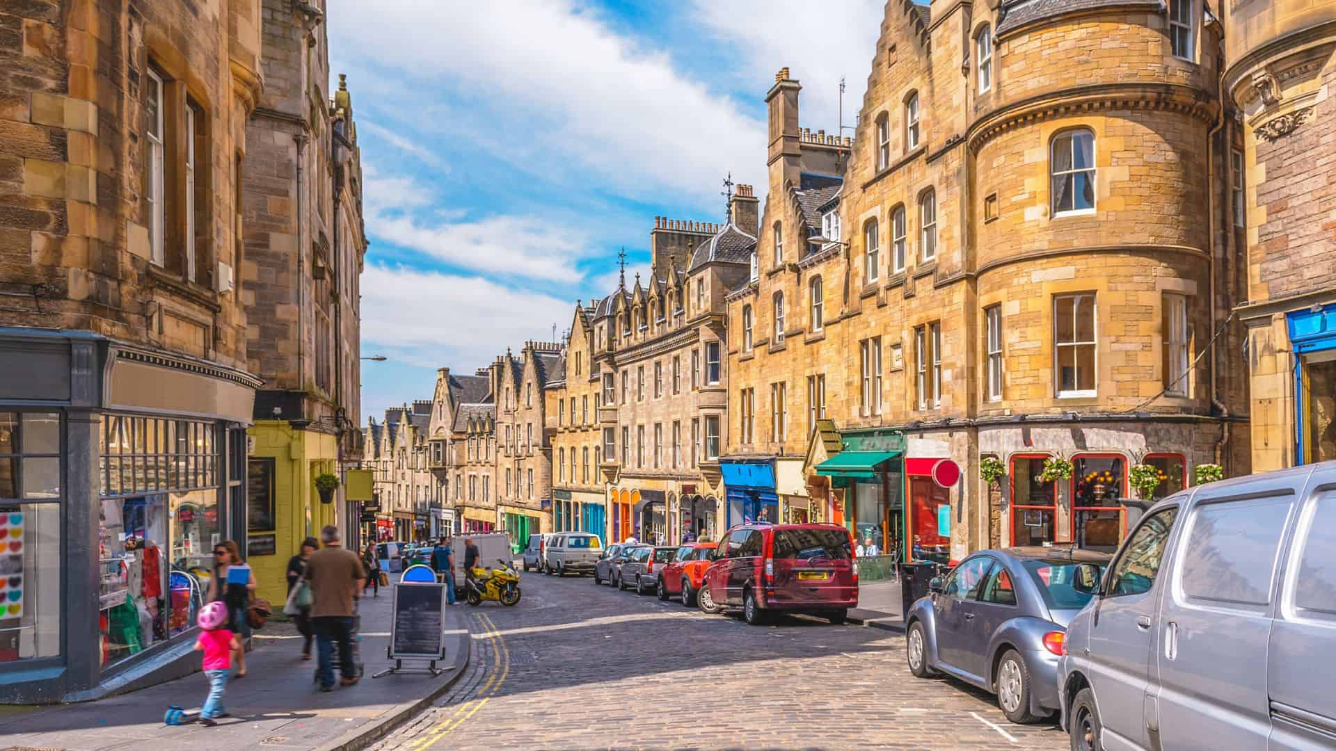 People walk along a cobblestone street lined with historic stone buildings and parked cars on a sunny day.