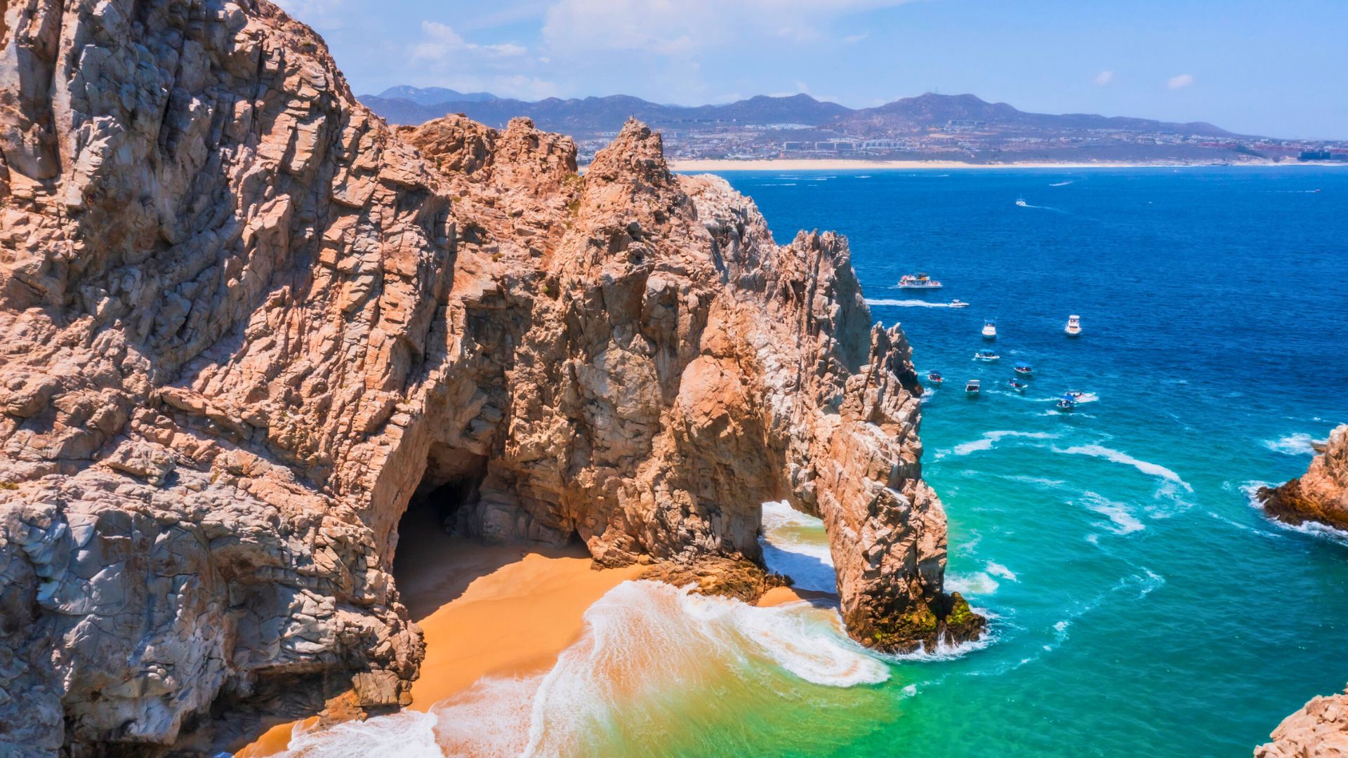 Rocky cliffs and sandy beach beside turquoise ocean, with boats on the water and mountains in the distance.