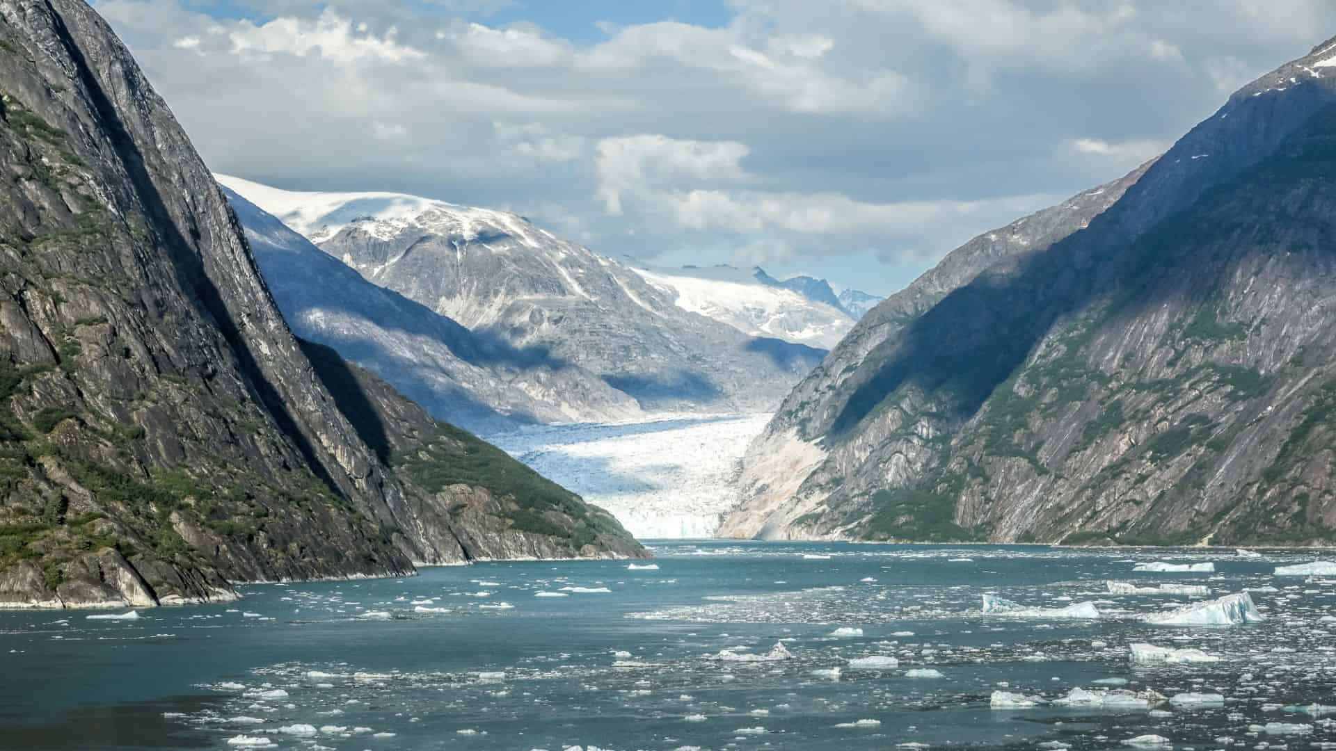 A glacier flows between steep mountain valleys, with ice floating on the water under a partly cloudy sky.