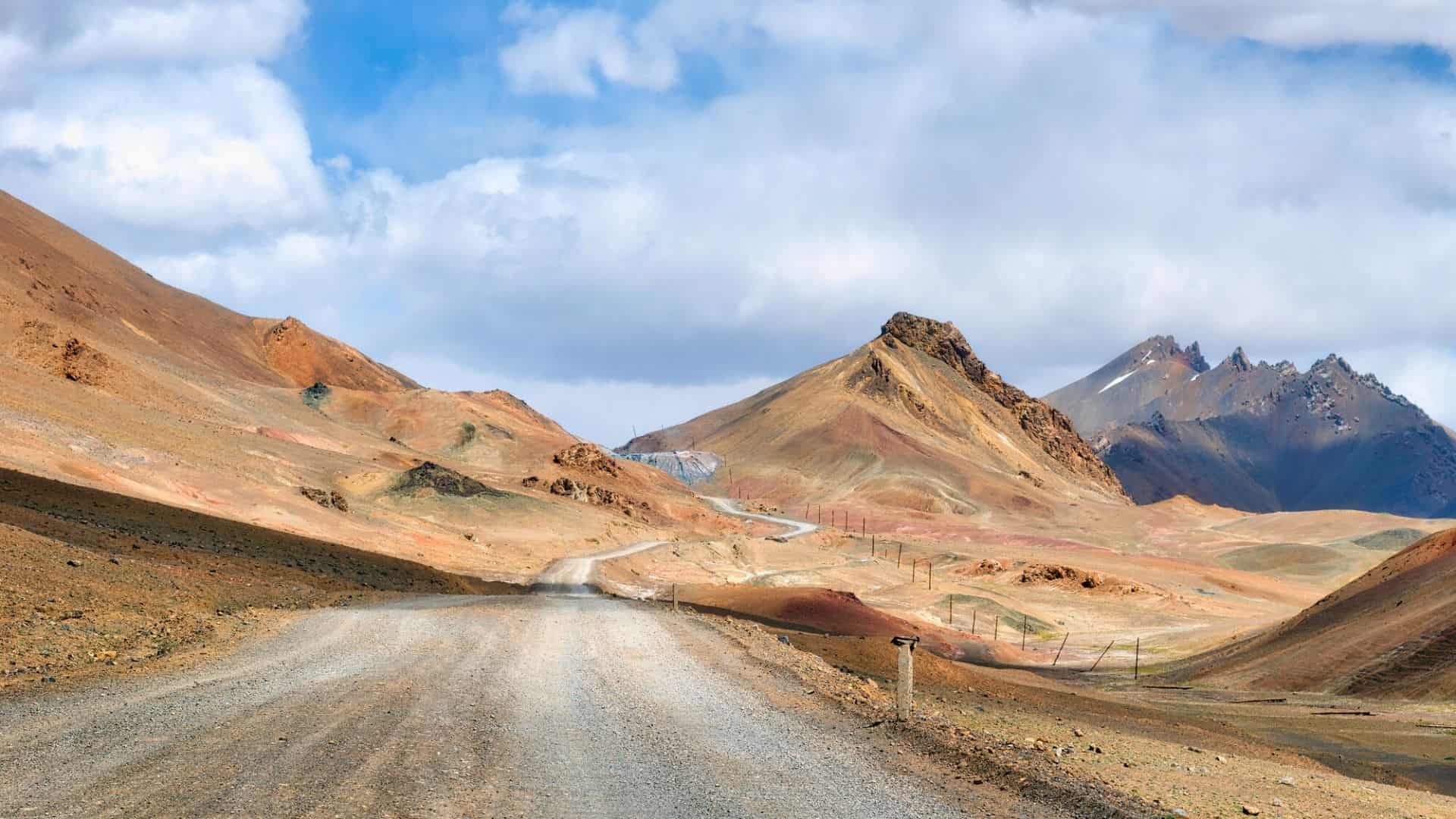 A winding dirt road stretches through barren, rocky mountains under a partly cloudy sky.
