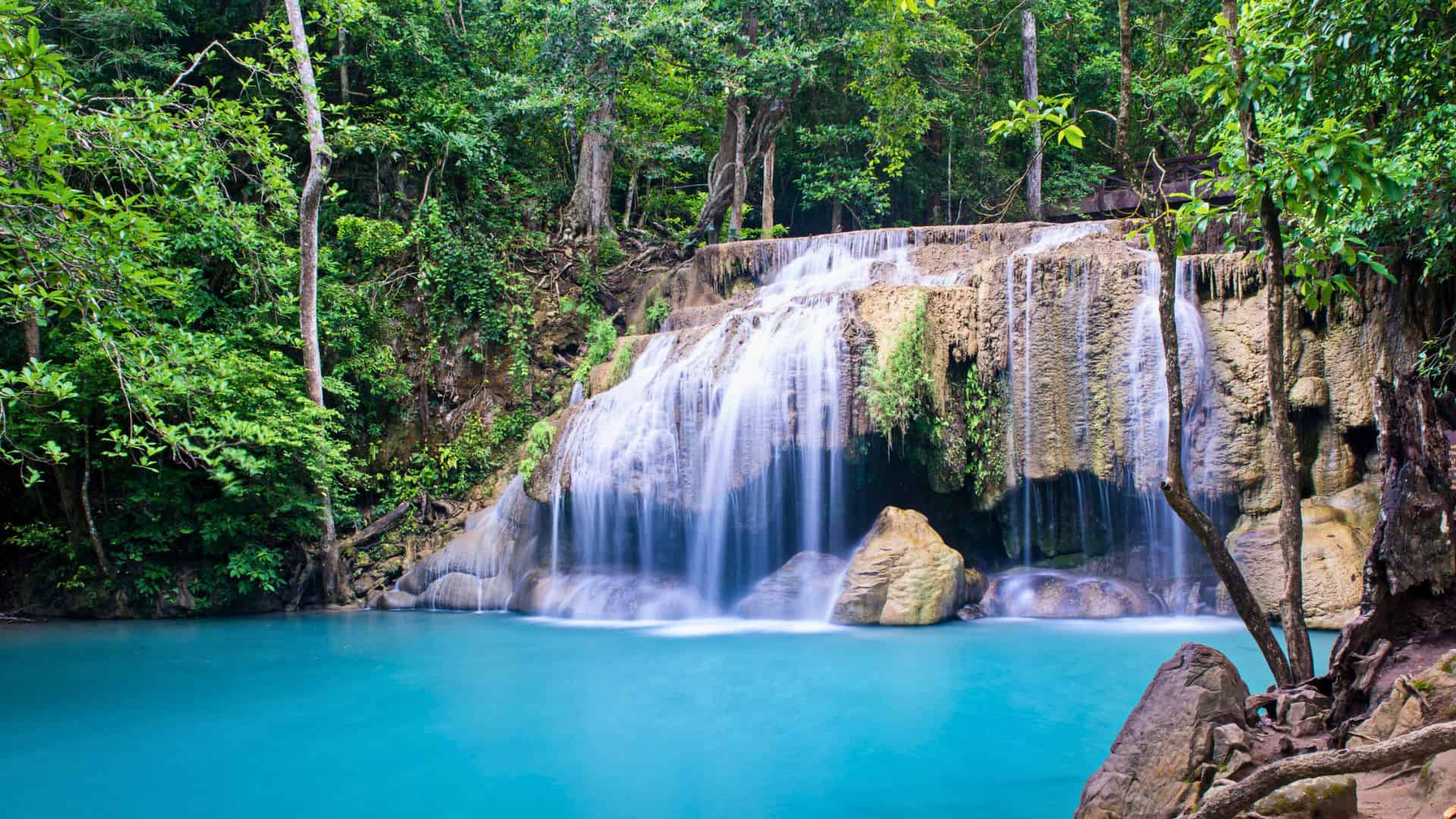 Waterfall cascading over rocks into a bright blue pool, surrounded by lush green forest.
