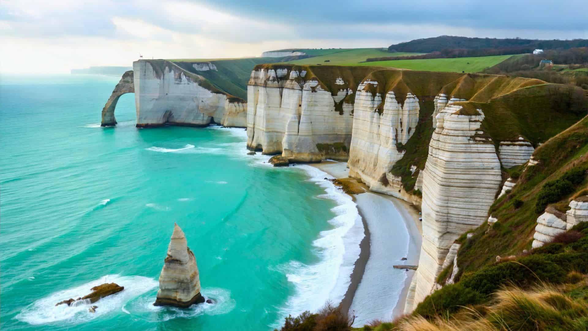 White chalk cliffs with natural arches rise above turquoise sea and sandy beaches under a cloudy sky.