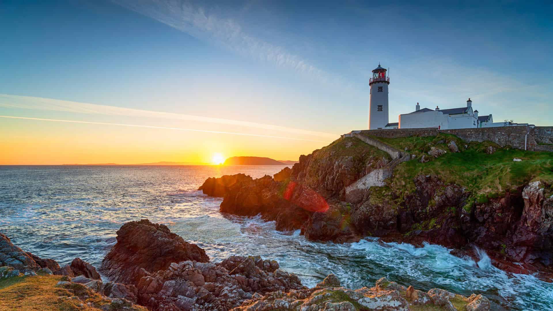 A lighthouse on a rocky coast at sunset with waves crashing and the sun low on the horizon.