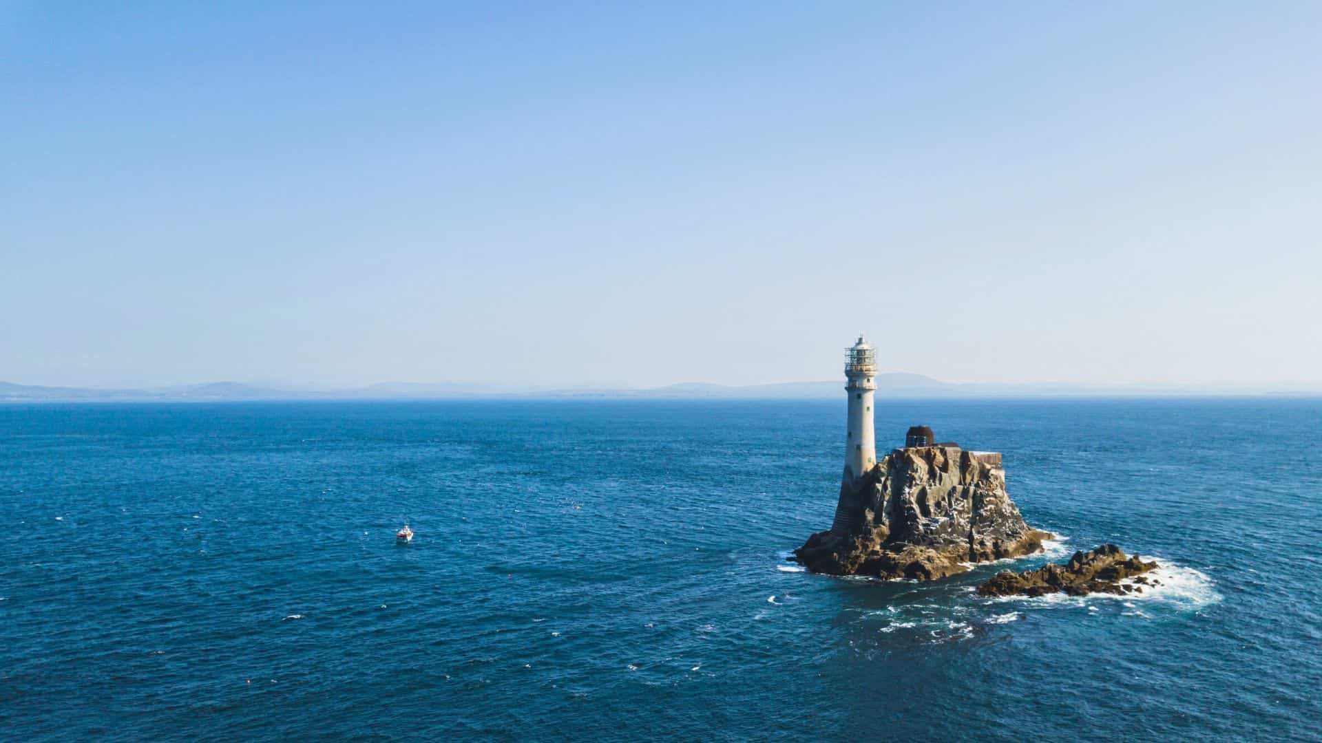 A solitary lighthouse stands on a rocky island surrounded by the blue ocean under a clear sky.