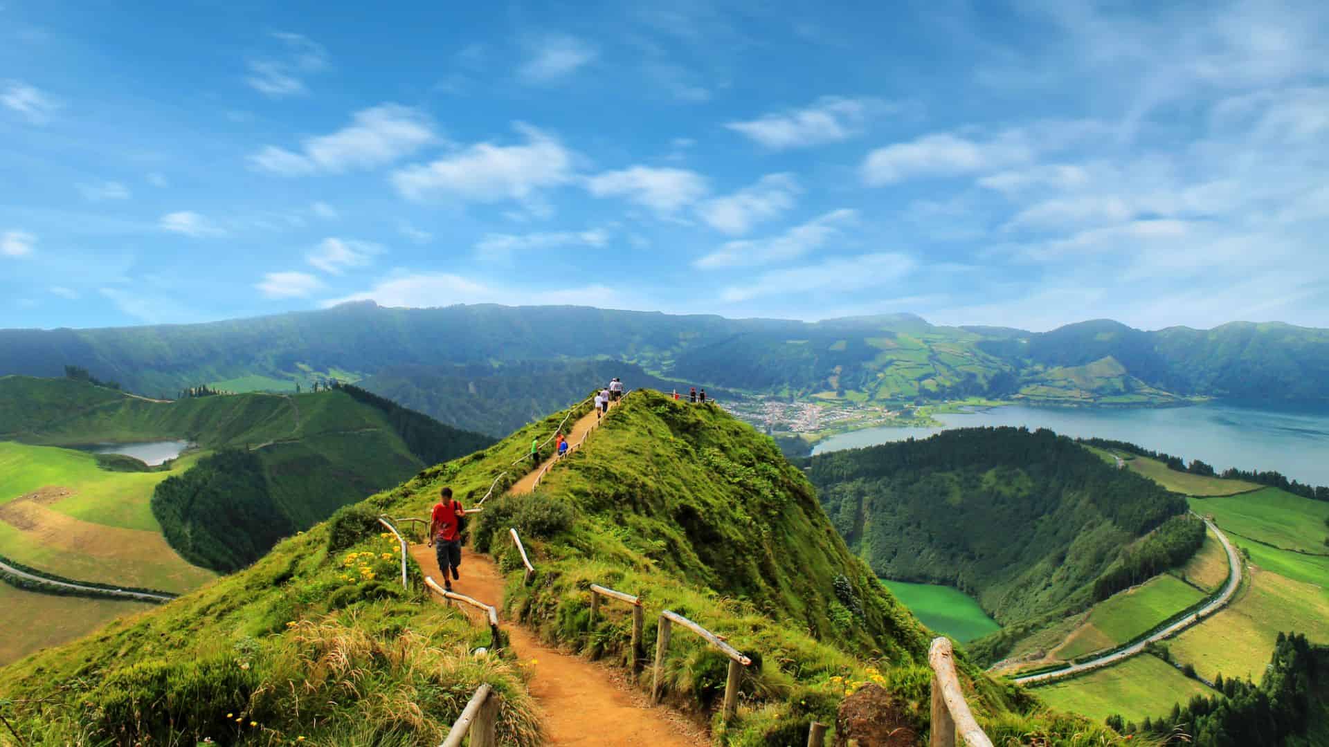 People hike along a scenic, green mountain ridge overlooking fields, lakes, and distant hills under a blue sky.
