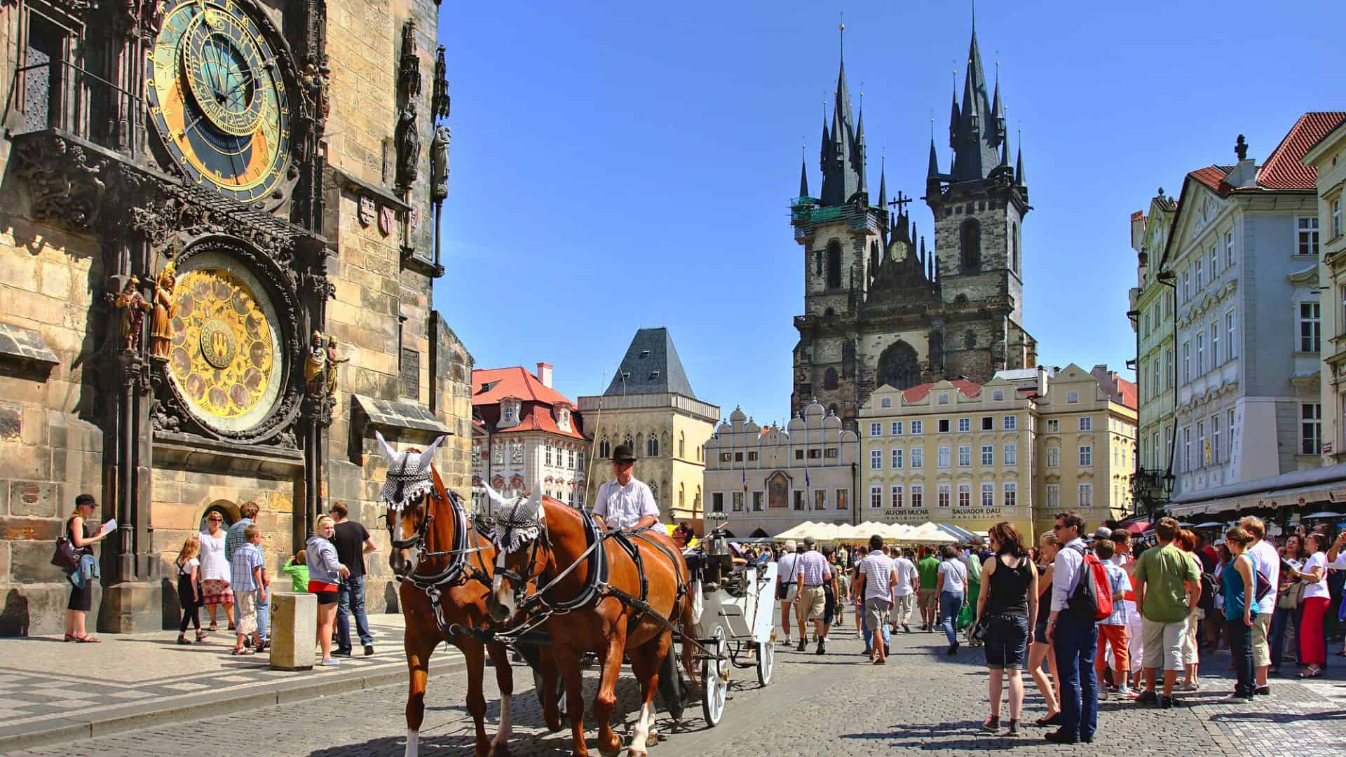 Horse-drawn carriage in a lively European square with an astronomical clock and gothic church in the background.