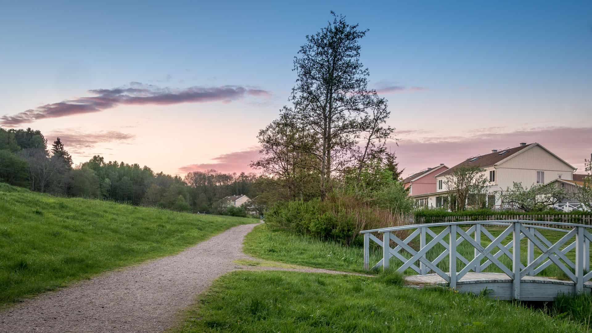 A gravel path leads past houses and over a small wooden bridge at sunset in a grassy, tree-lined area.