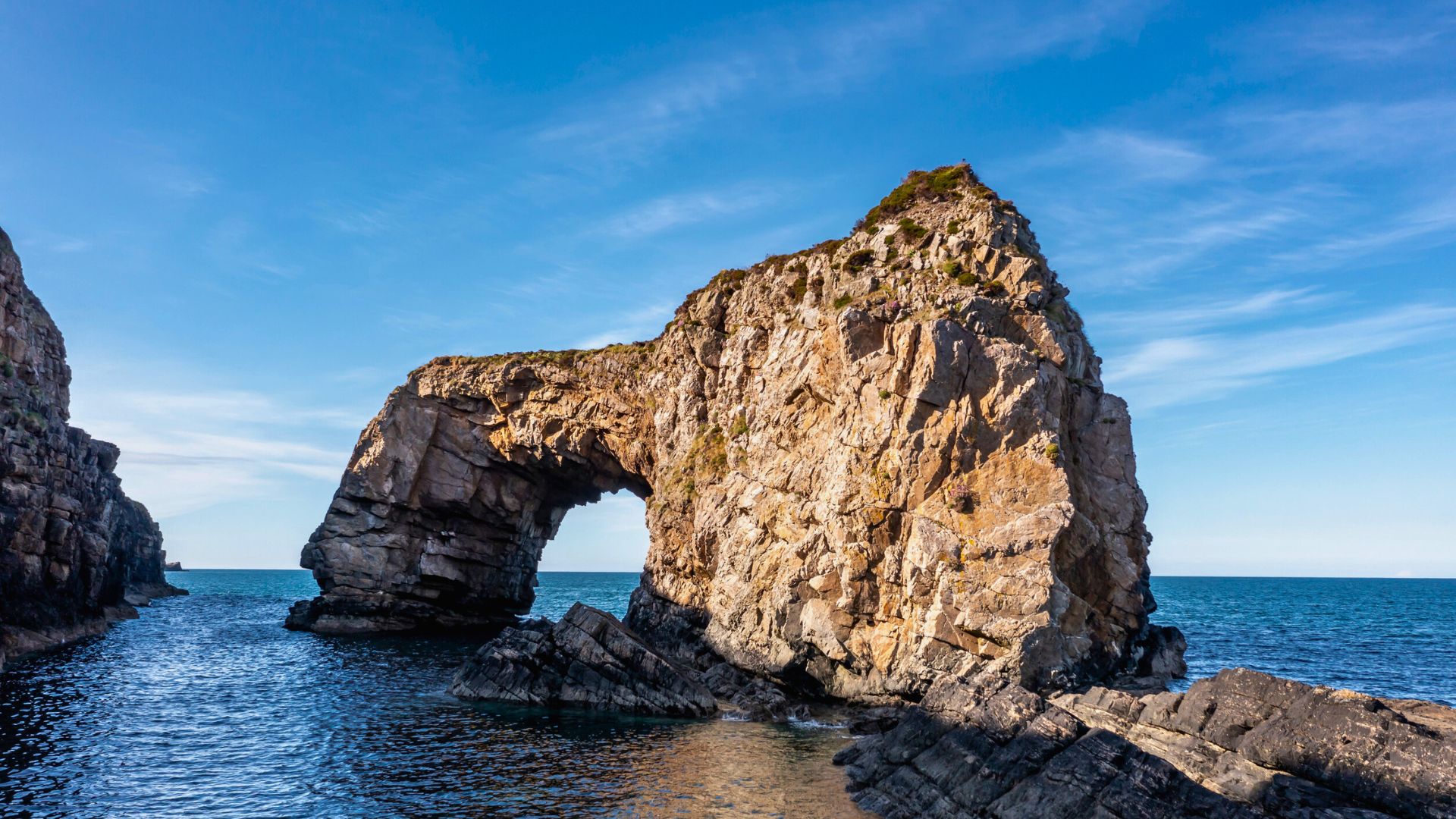 Large rocky sea arch rises from the blue water under a clear sky, with some greenery on top.
