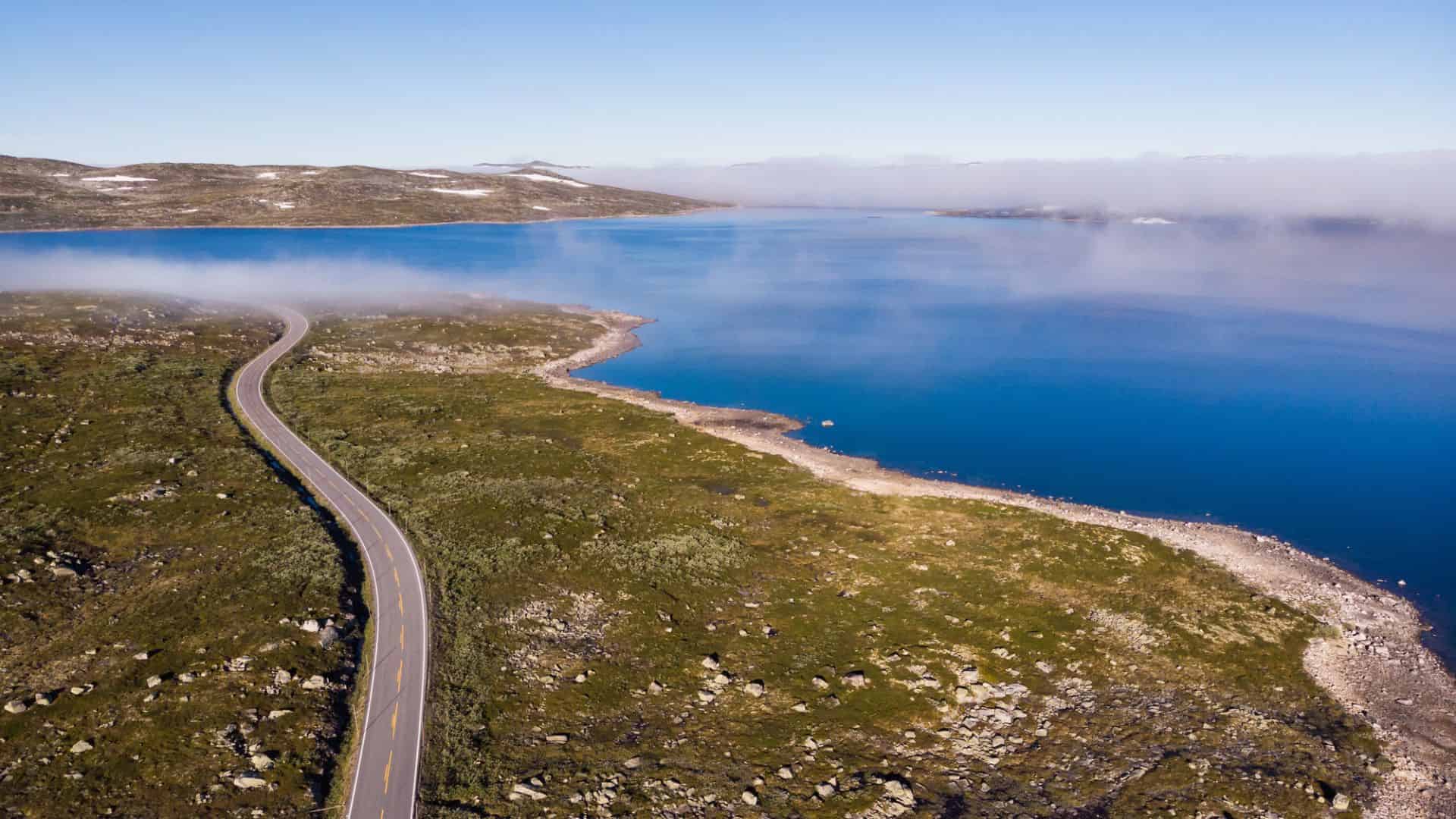 A winding road runs along a calm blue lake with grassy hills and light fog in the distance.
