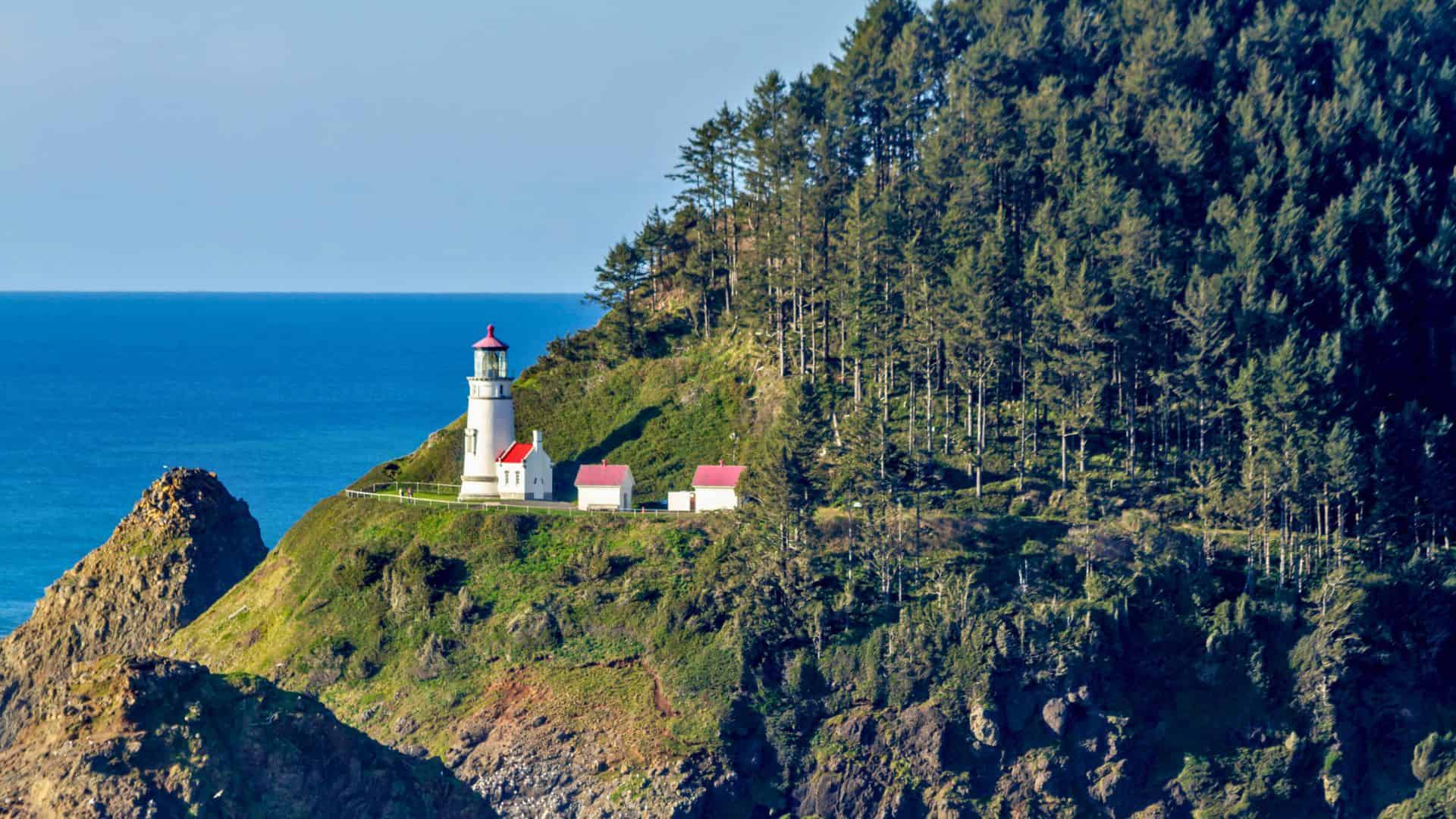 White lighthouse with red roof sits on a green, forested cliff overlooking the blue ocean under a clear sky.
