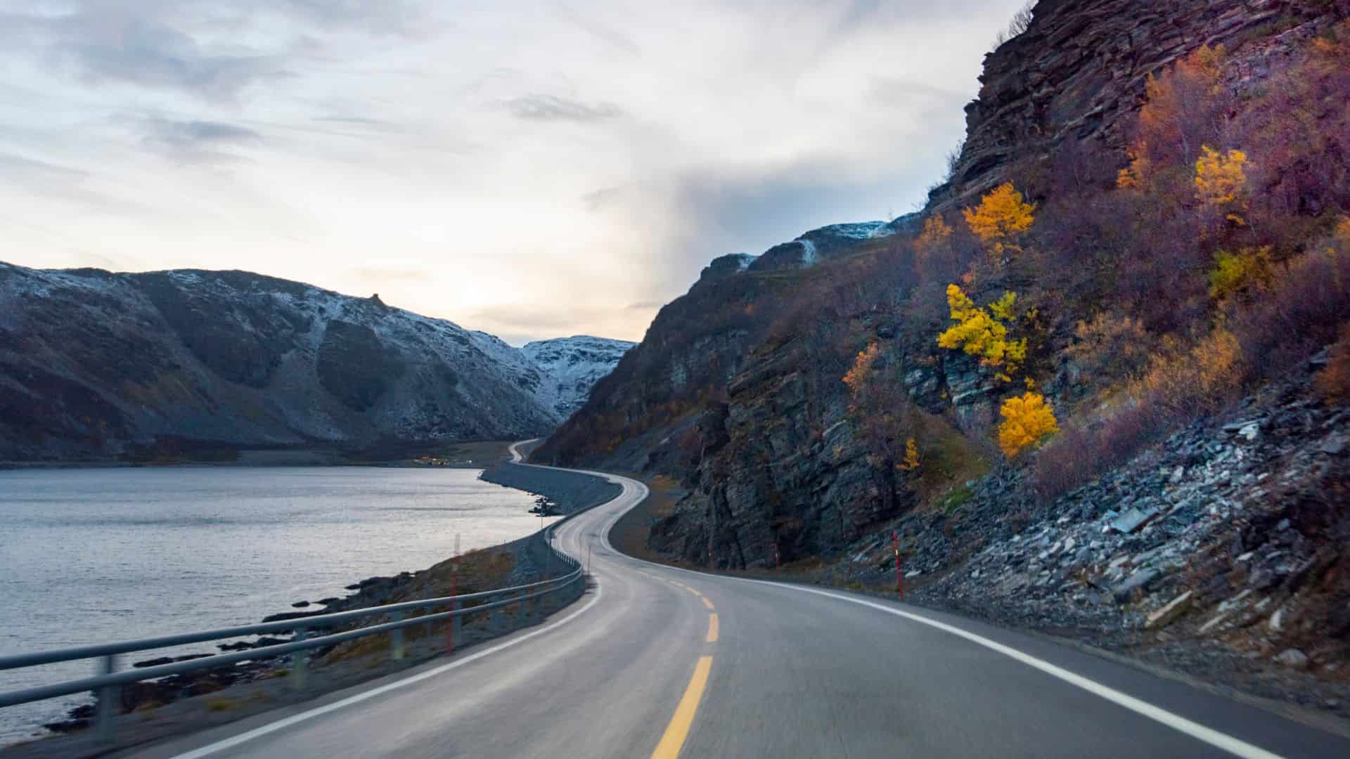 Curved road along a rocky coastline, with autumn trees and snow-capped mountains under a cloudy sky.